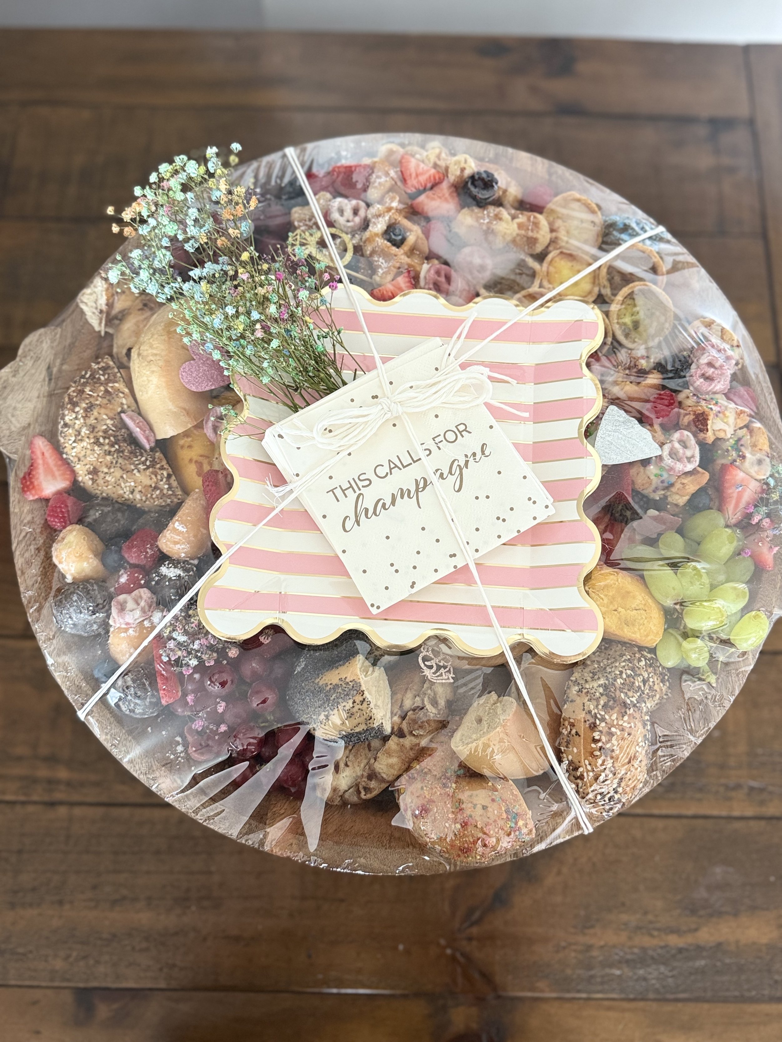 A round charcuterie board filled with an assortment of cookies, candies, and fruit, wrapped in clear plastic, with a pink and white striped decorative tag attached with white string created by Cannon's Table.