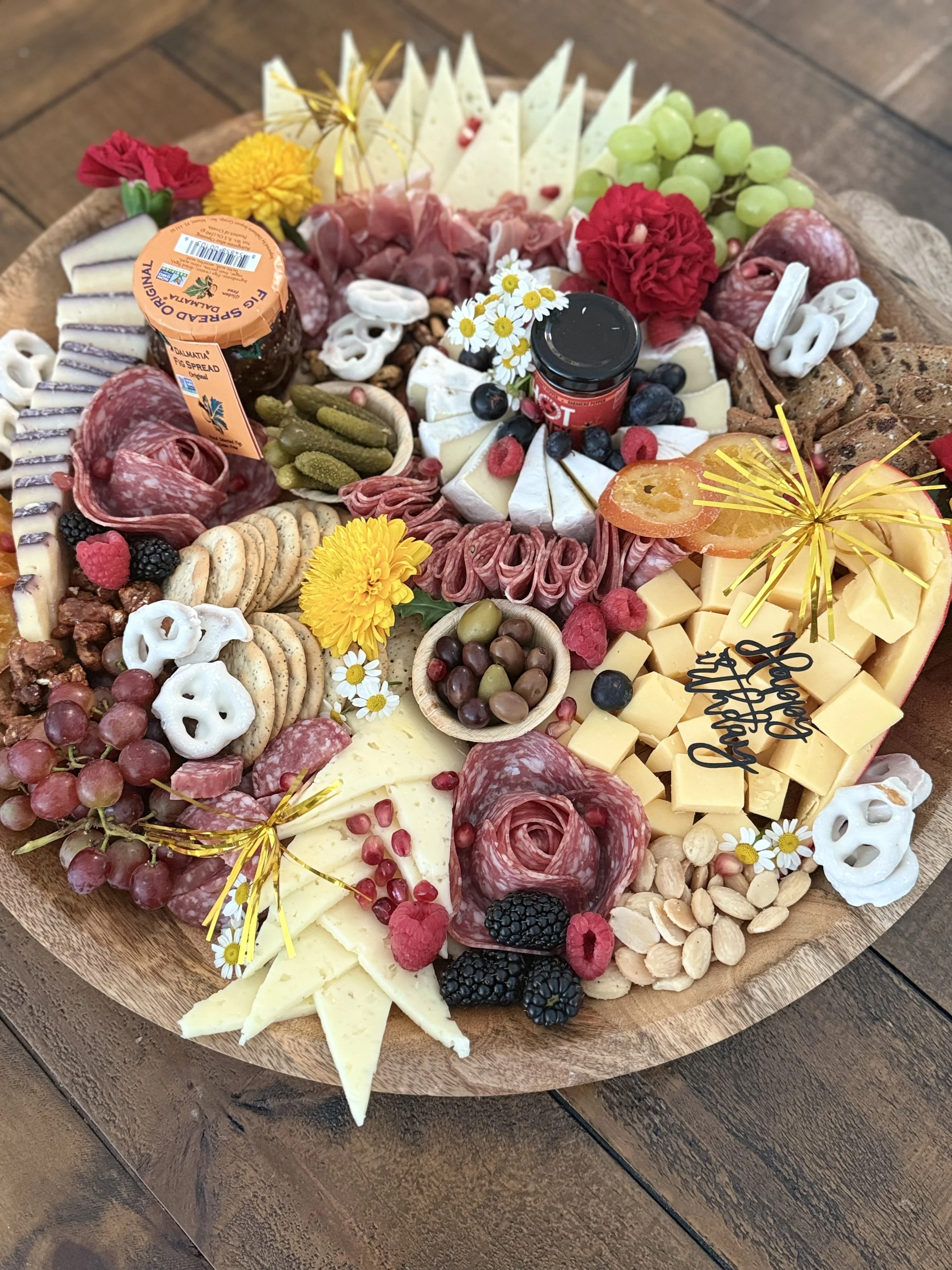 A large cheese and charcuterie board with various types of cheeses, crackers, cured meats, fresh and dried fruits, nuts, pickles, olives, and decorative flowers, arranged on a wooden serving platter created by Cannon's Table.