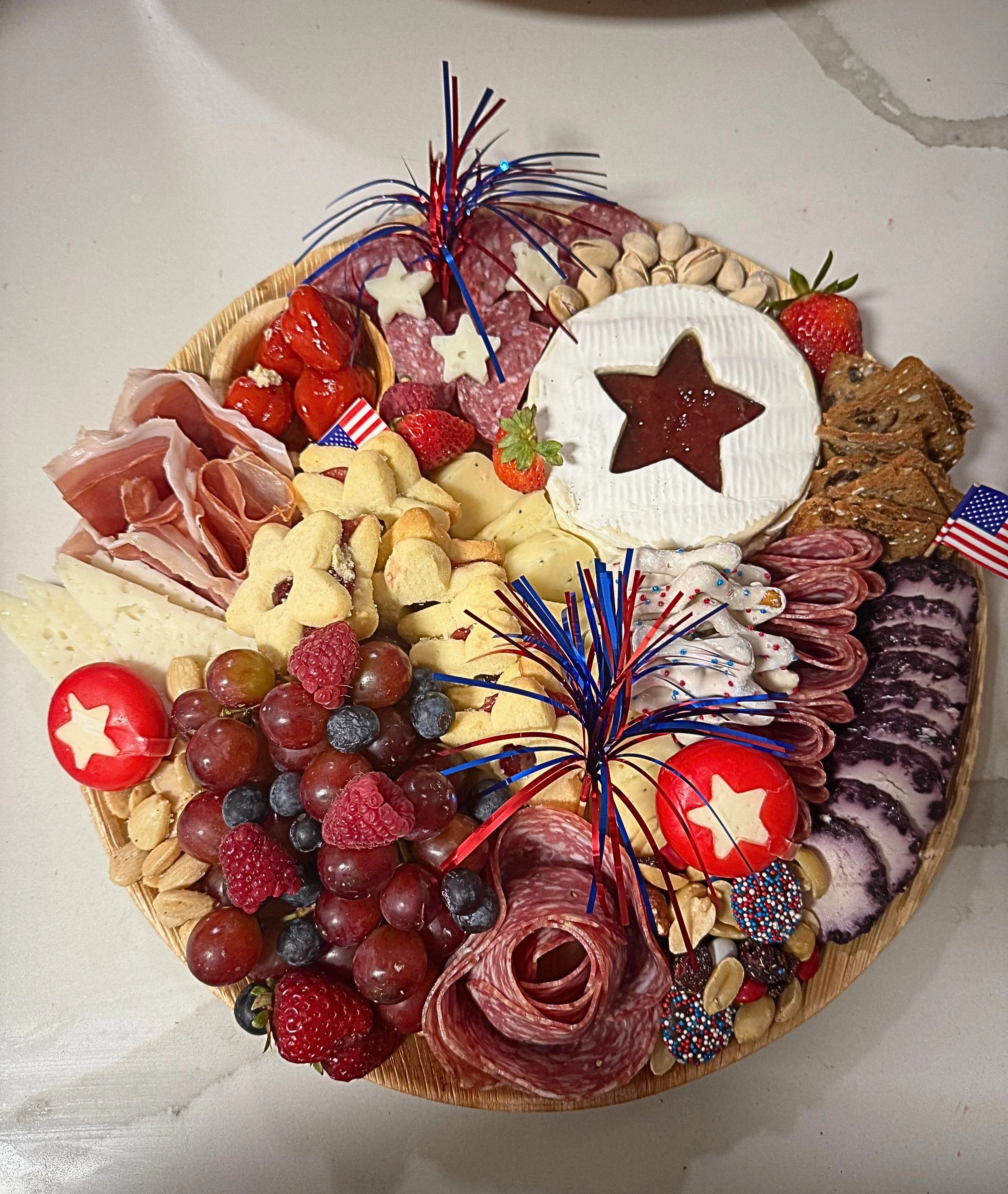 A round charcuterie board with assorted meats, cheeses, fruits, nuts, and decorated with red, white, and blue firework picks, created by Cannon's Table.