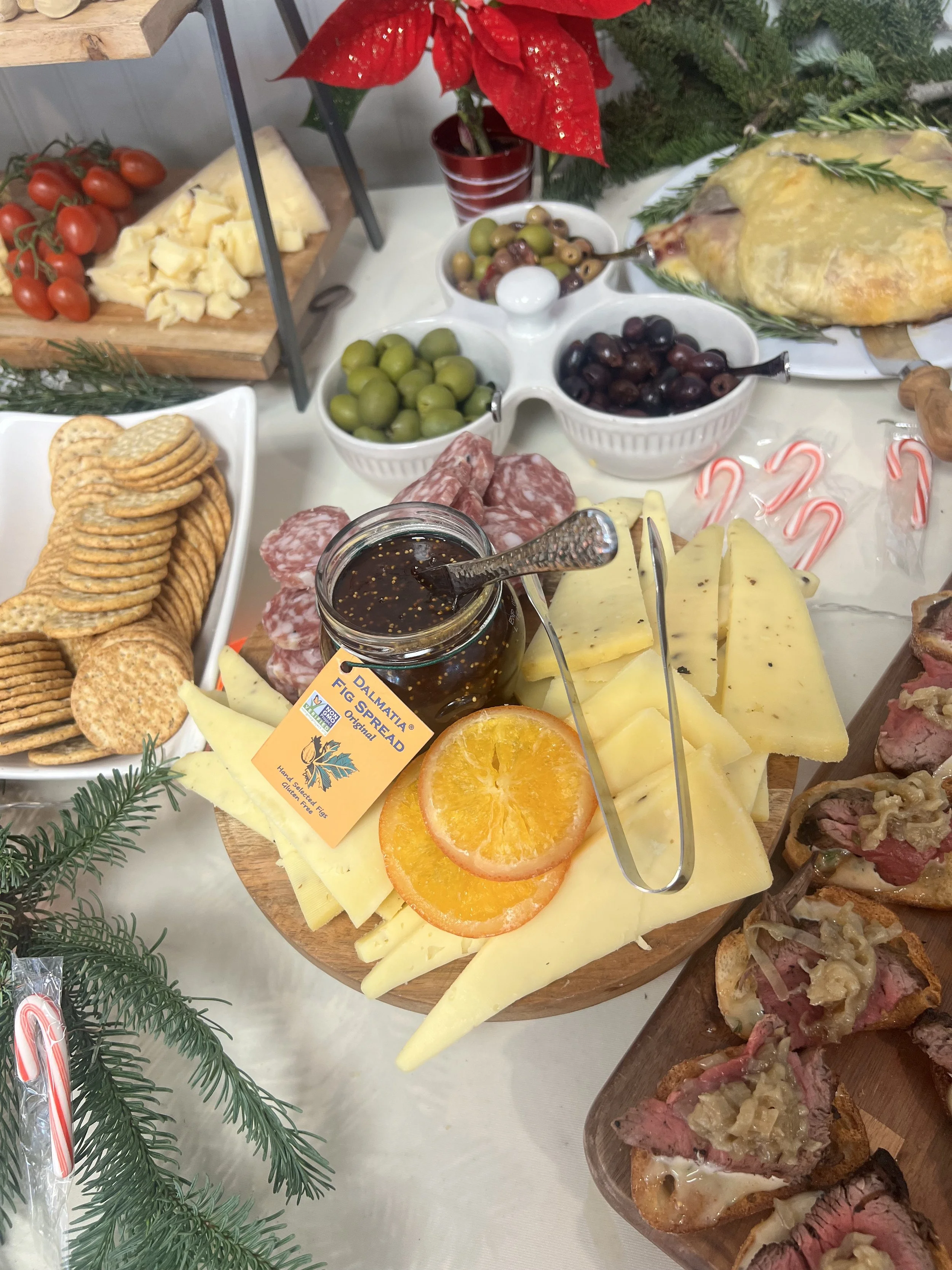 A cheese platter with sliced cheese, orange slices, a small jar of fig spread, crackers, salami, and various bowls of olives and other snacks, created by Cannon's Table.