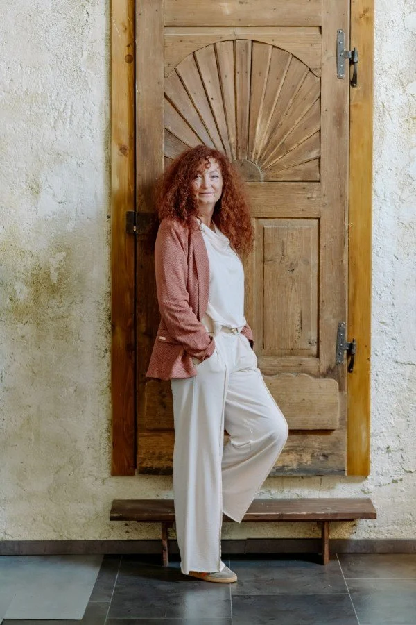Renate Heimerl steht seitlich an eine antike Holztür gelehnt. Sie trägt eine helle weite Hose und ein gleichfarbiges Top. Darüber einen Blazer in altrosa. Ihre Hände sind in den Hosentaschen.