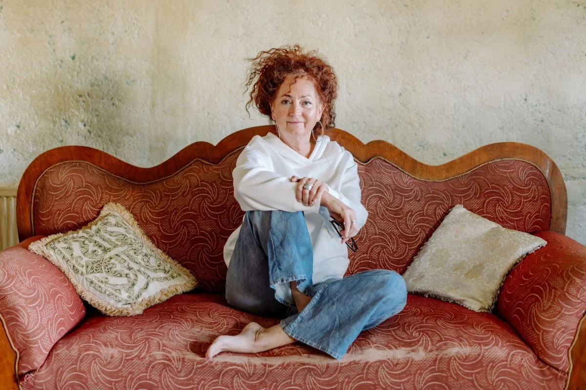 Eine Frau mit lockigem Haar sitzt entspannt auf einem antiken Sofa. Sie trägt ein weißes Sweatshirt und blaue Jeans mit einer Hand auf ihrem Bein hält sie eine Brille. Der Hintergrund zeigt eine grob verputze helle Wand.