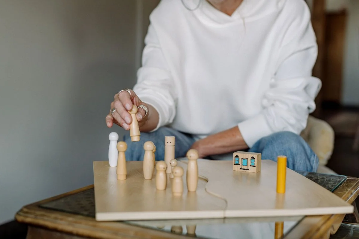 Ein Holzbrett für systemische Aufstellungsarbeit steht auf einem Tisch. darauf sind verschiedene Holzfiguren, die als Vertreter in der Aufstellung fungieren. Renate Heimerl sitzt im Hintergrund und hält eine der Figuren in der Hand.