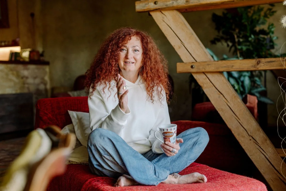 Renate Heimerl sitzt mit gekreuzten Beinen auf einem roten Sofa. In einer Hand hält sie eine Kaffeetasse mit der anderen macht sie eine Geste, als würde sie zum Zuschauer direkt sprechen.