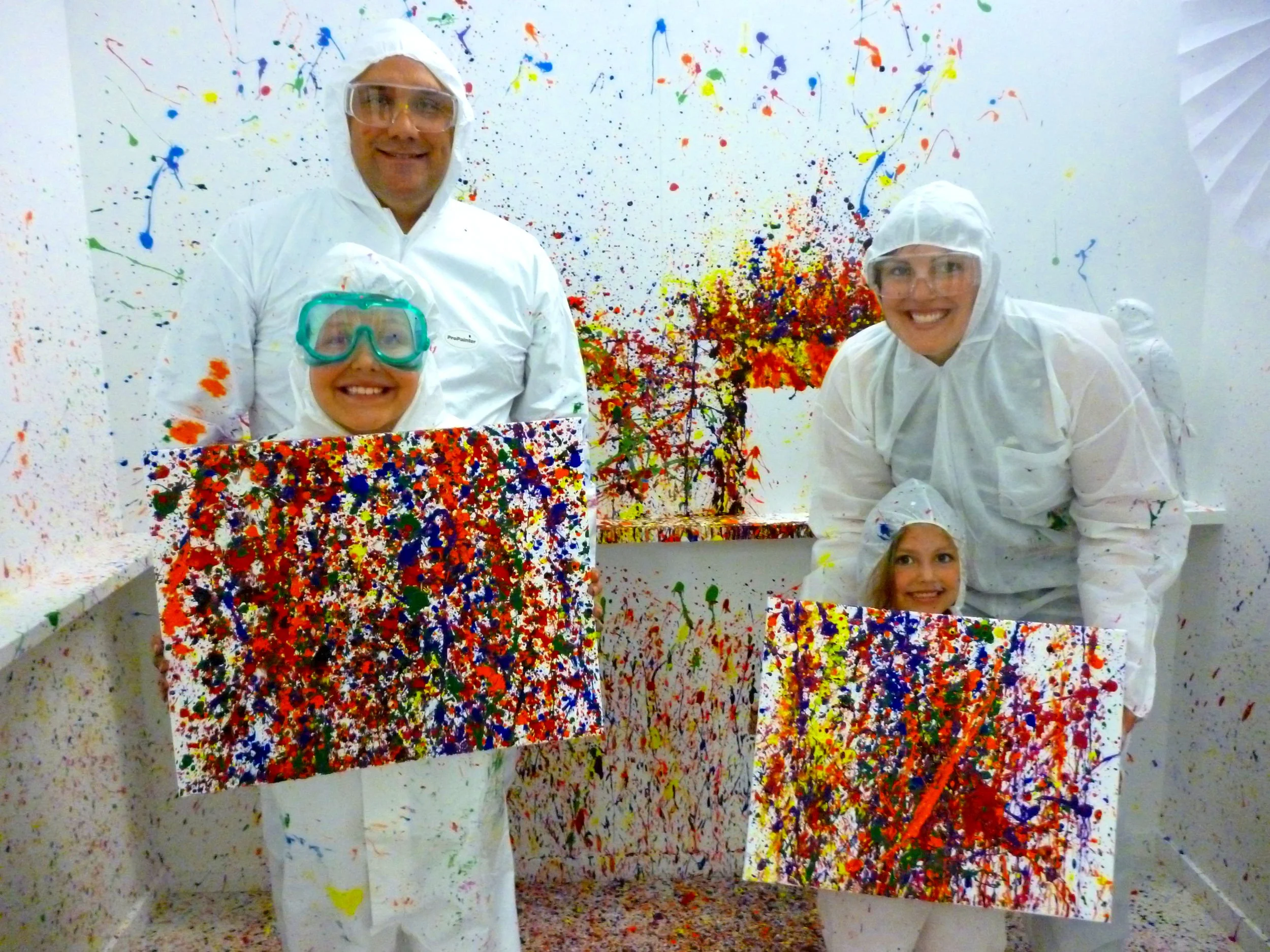 Four people in white protective suits and glasses, smiling, holding colorful splattered canvases, inside a room with walls splattered with colorful paint, indicating a fun painting activity.
