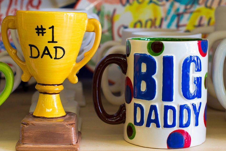Colorful ceramic mug with polka dots and the text 'BIG DADDY' next to a yellow trophy mug with the text '#1 DAD' on it, both displayed on a shelf at Wet Paint Pottery Saskatoon.
