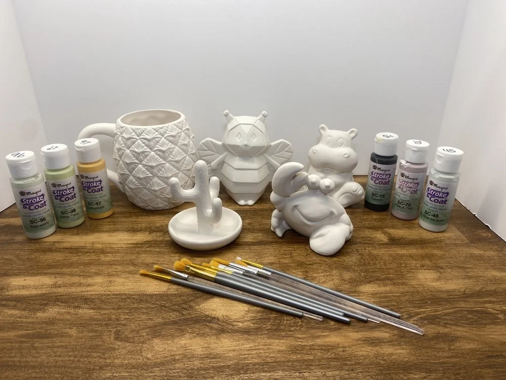 Several paint bottles, paintbrushes, and ceramic decorative figures on a wooden surface.