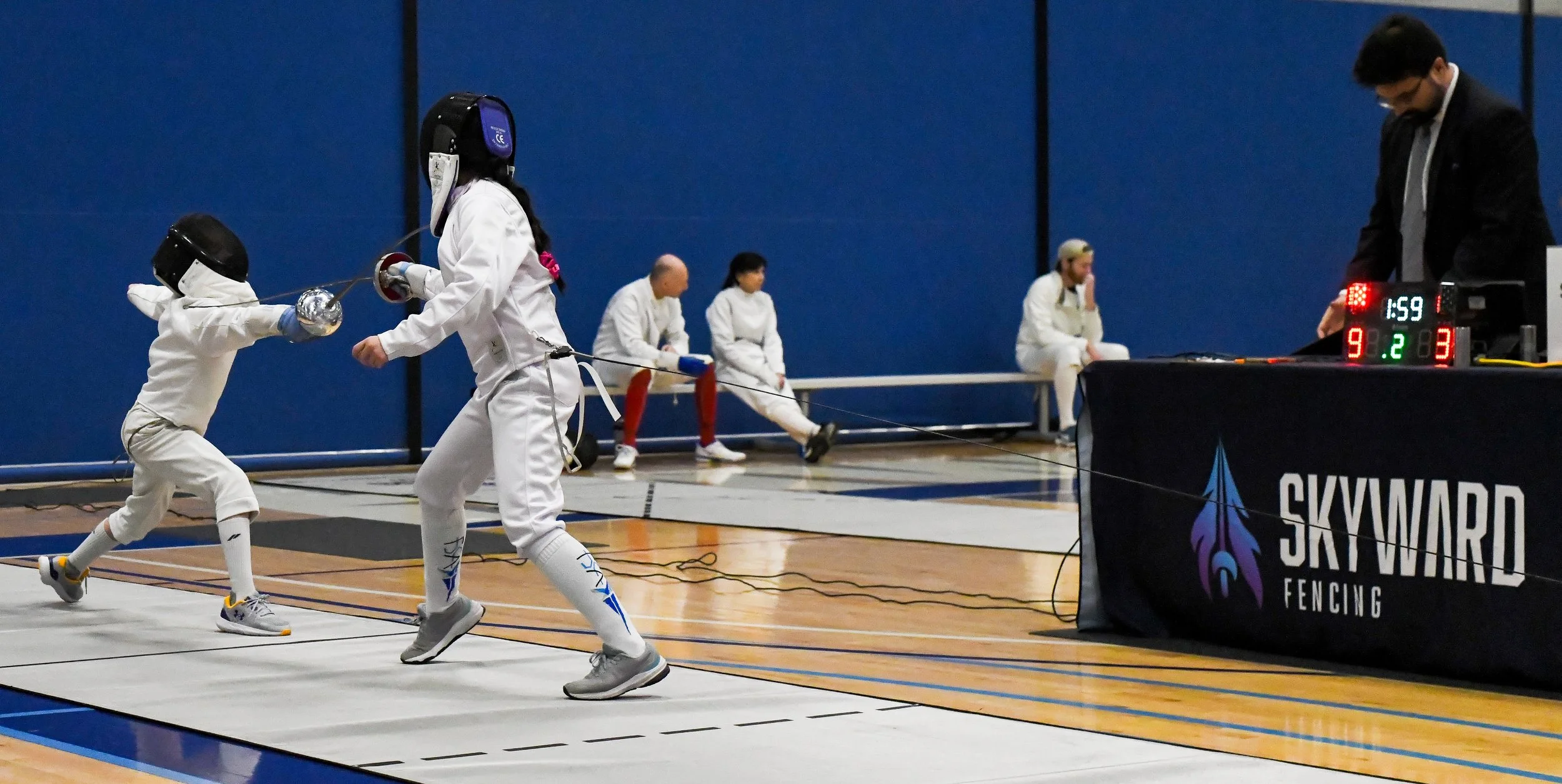 Two fencers dressed in white fencing gear engaged in a fencing match, with a digital scoreboard displaying scores and the logo of Skyward Fencing in the foreground. Two individuals are seated in the background against a blue wall.