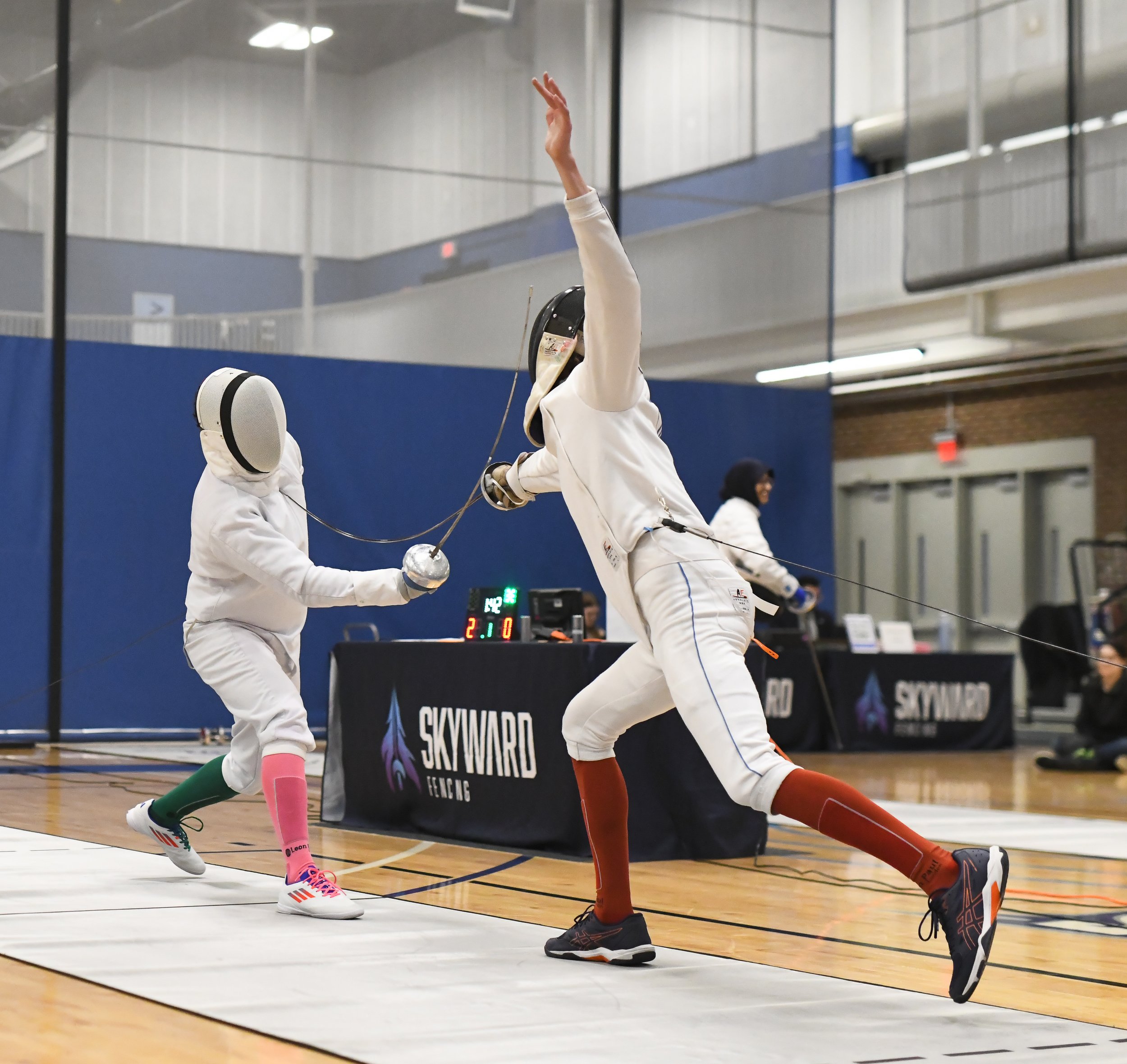 Two female fencers competing in a fencing match indoors. They are dressed in fencing gear, including masks, jackets, and gloves. The fencer on the right is extending her arm with her foil, while the other appears to be preparing to respond. The background includes a digital scoreboard and a fencing piste with barriers.