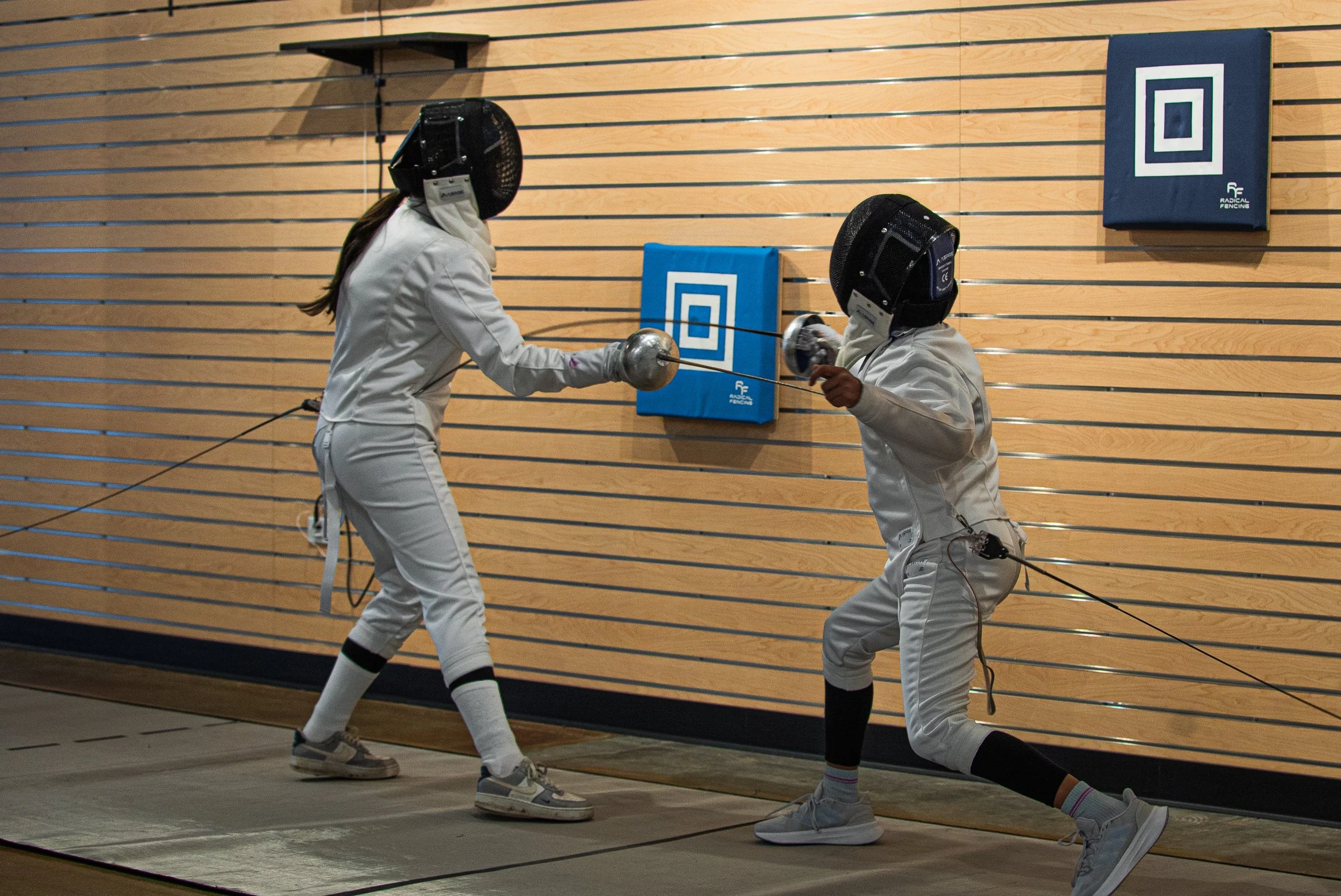 Two fencers practicing foil fencing indoors on a wooden wall with padded mats. One fencer is lunging forward, the other is parrying with their foil.