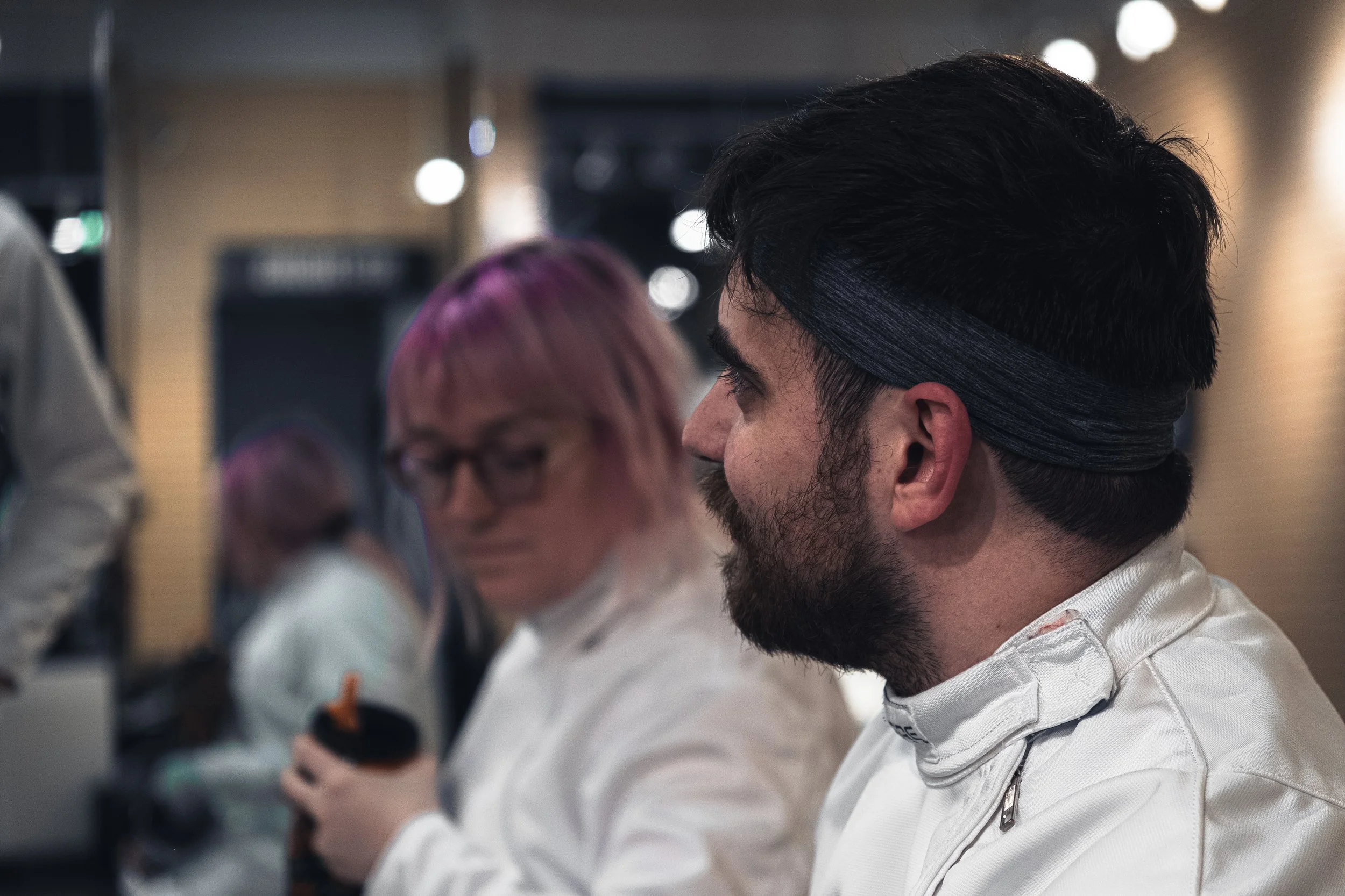 Close-up of a man with dark hair, beard, and a headband, wearing a white chef's coat, working at a kitchen counter with other people in the background.