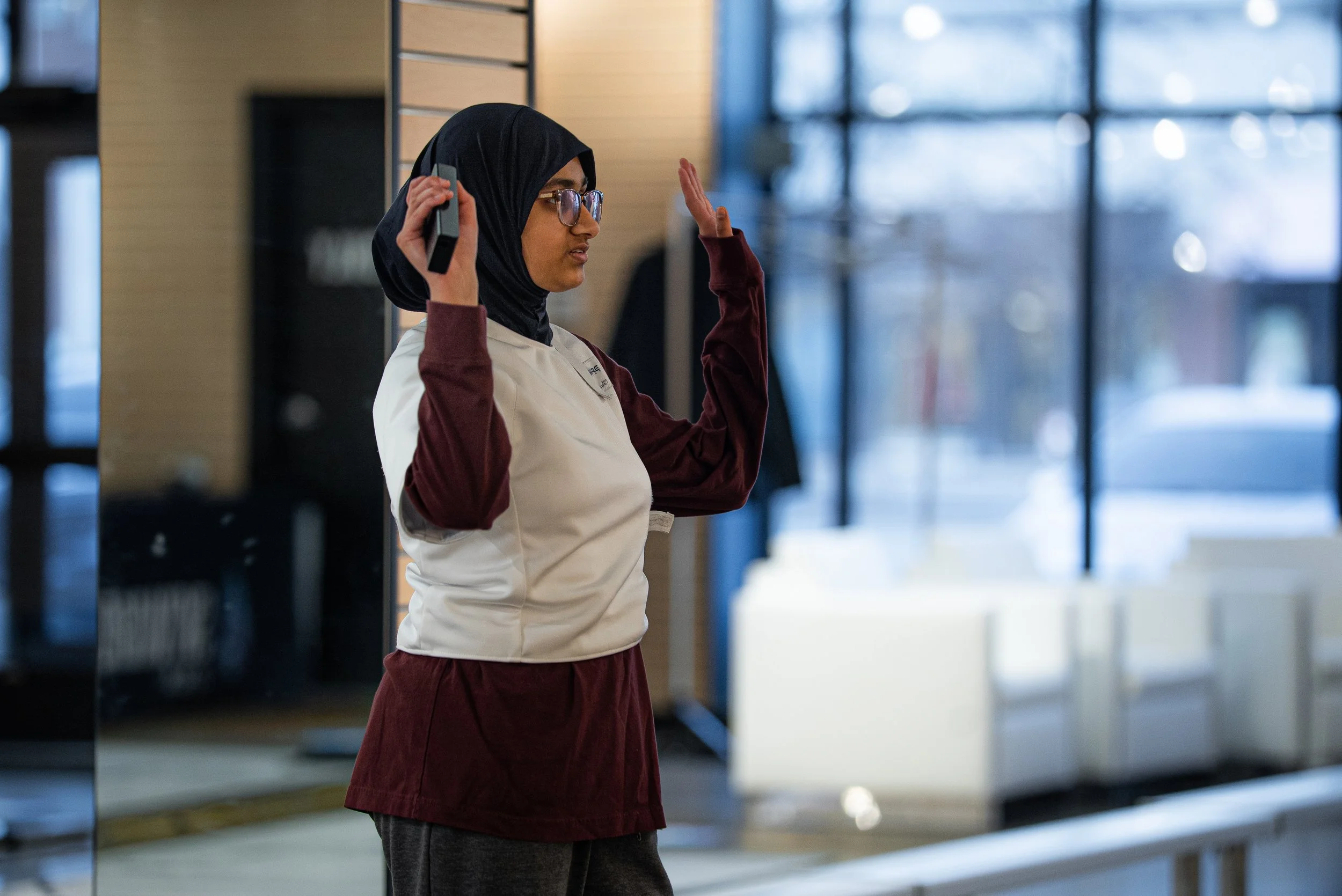 A young woman with glasses and a black hijab, wearing a maroon and white jacket, is holding a cellphone to her ear and gesturing with her left hand inside a modern building with large windows and blue accents.