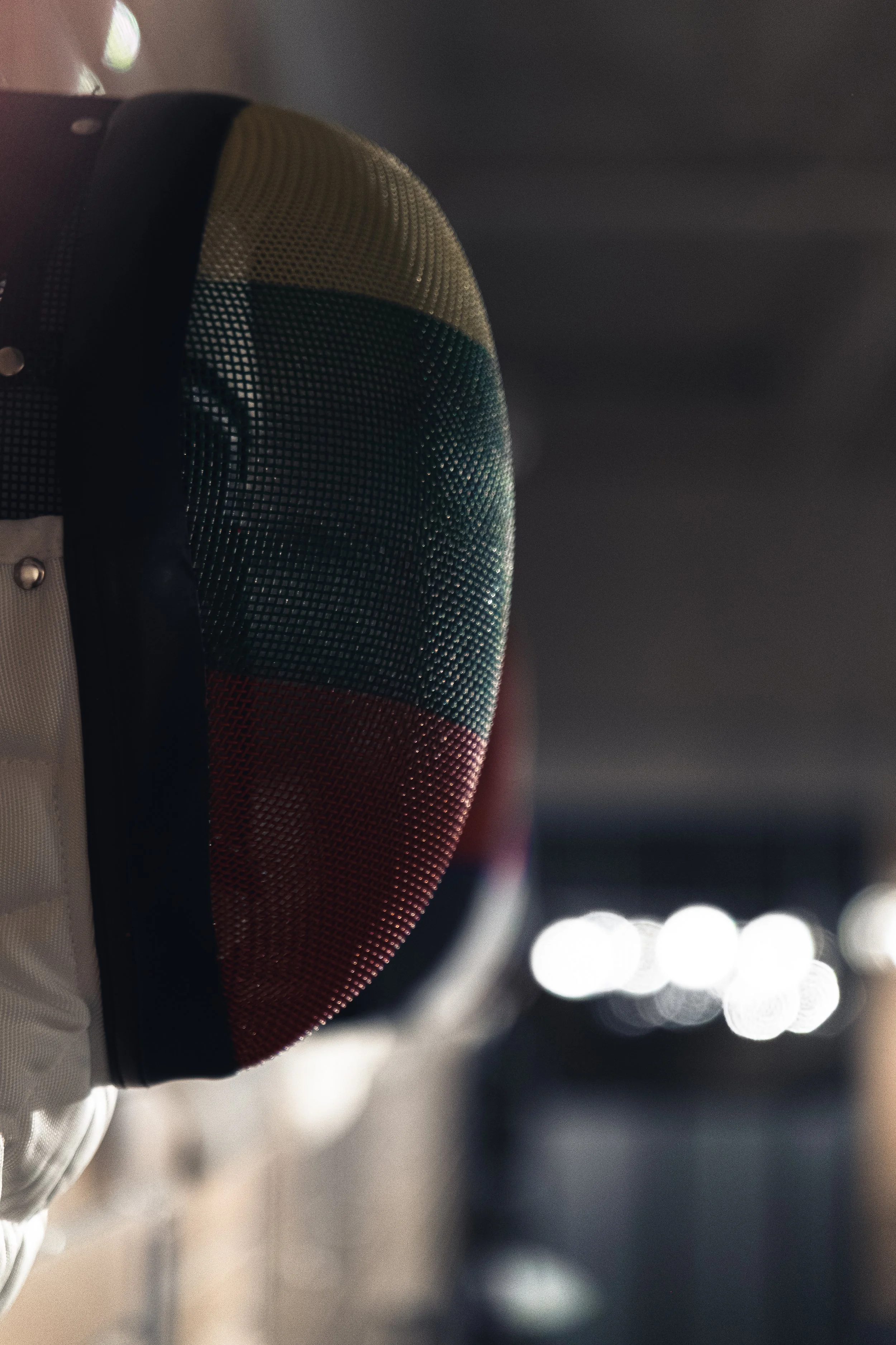 Close-up of a fencing helmet with a metallic mesh faceguard, showing reflections of lights on the mesh, set against a dark background with blurred bright lights.