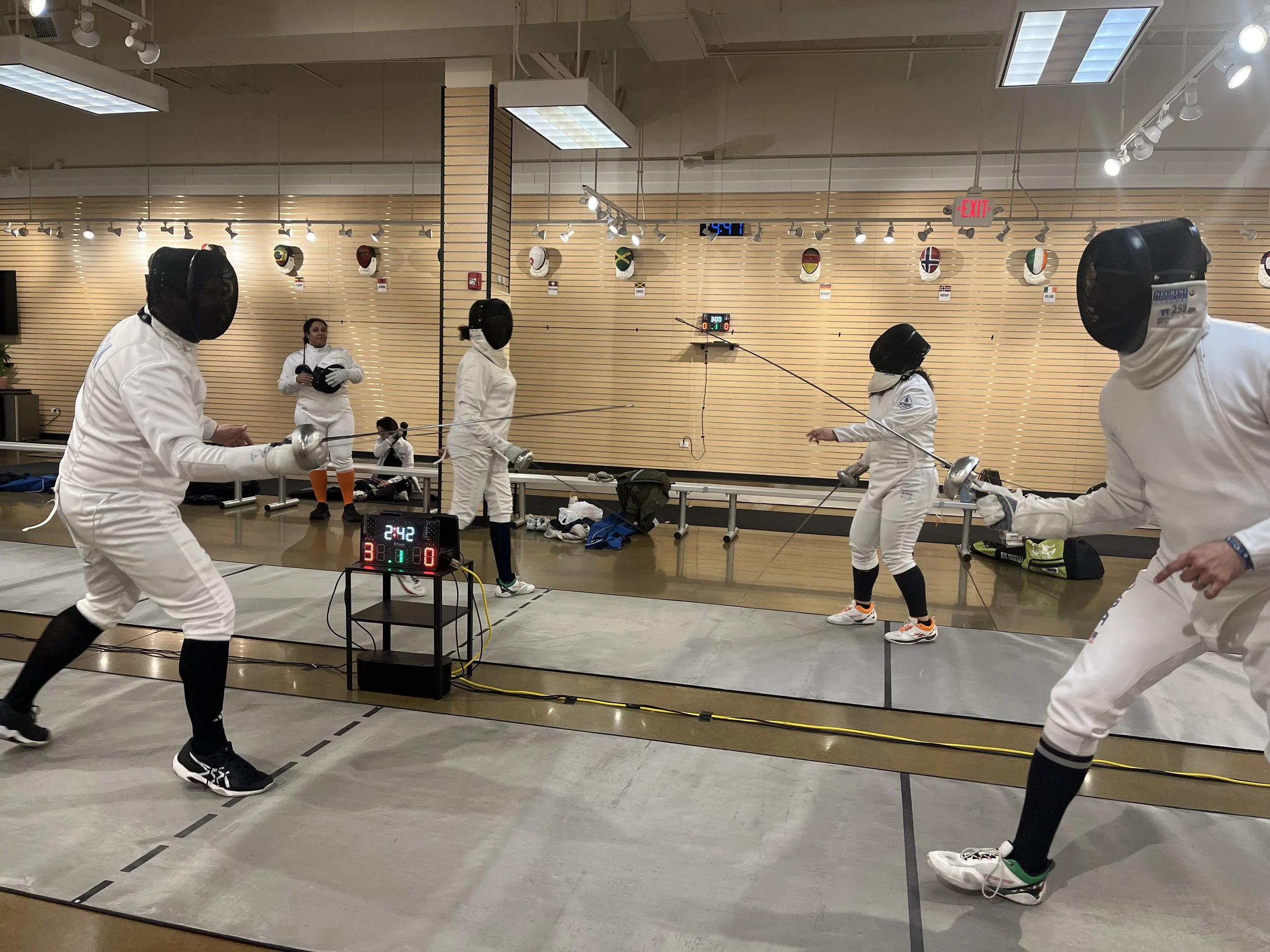 Fencing match with fencers in white jackets and masks competing indoors, digital scoreboard showing current score, referees and spectators in background, decorative masks on the wall.