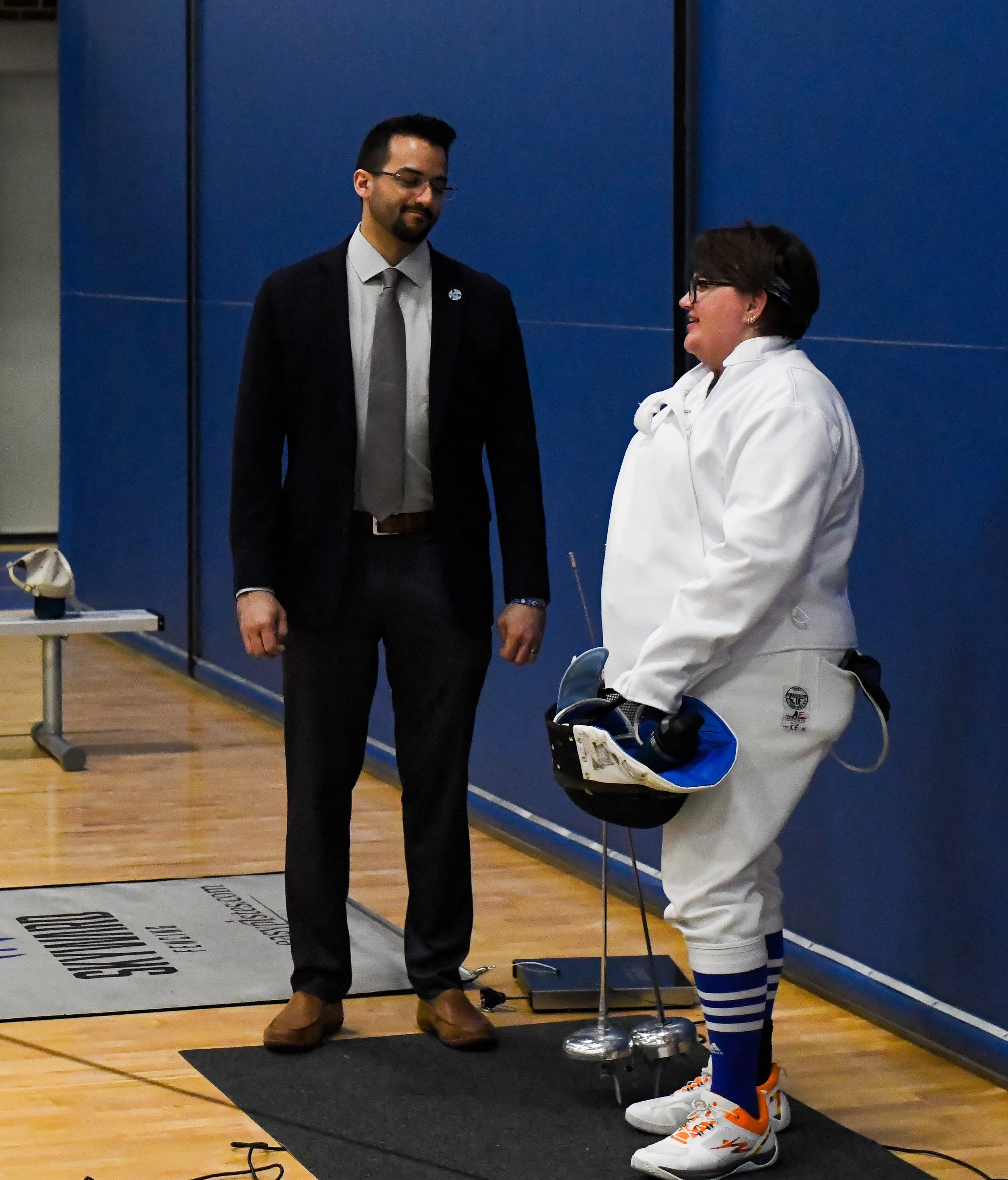 A man in formal attire standing and smiling at a young woman in a hockey uniform who is holding a hockey helmet and a trophy, inside a gymnasium with blue wall and wooden floor.