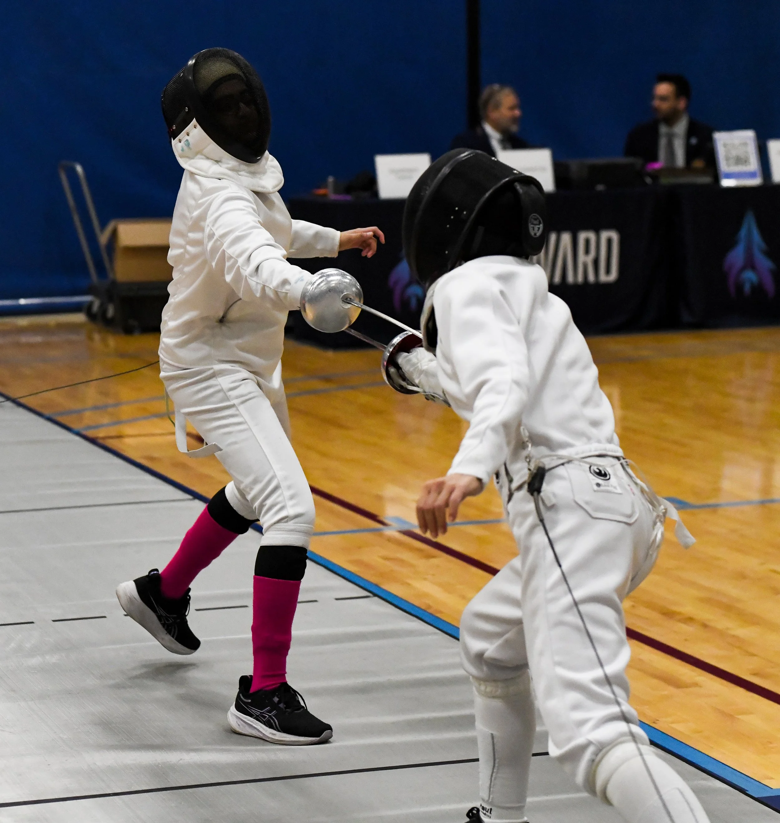Two fencers wearing white protective gear and helmets, engaged in a fencing match in an indoor gymnasium. One fencer is lunging forward with a foil, while the other is on guard. Two judges or officials are seated at a table in the background.