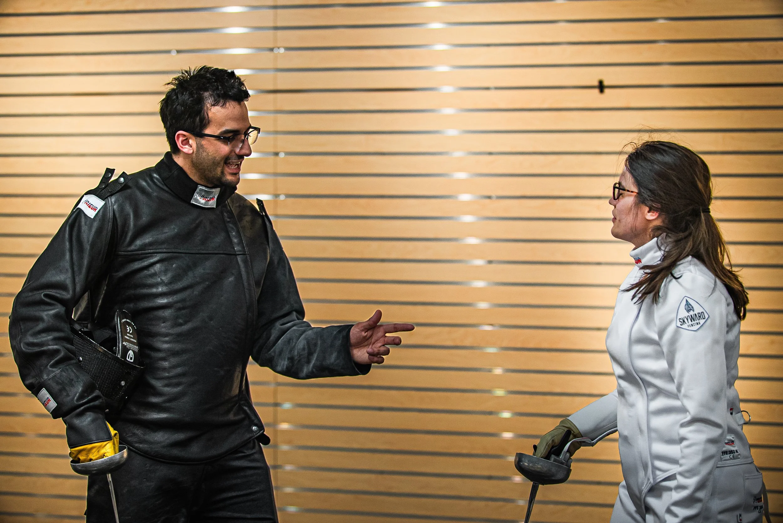 A man and a woman in racing gear are talking in front of a wooden wall. The man is wearing a black leather racing suit with patches, glasses, and has a crutch in his left hand. The woman is dressed in a white racing suit and glasses, holding a racing helmet.