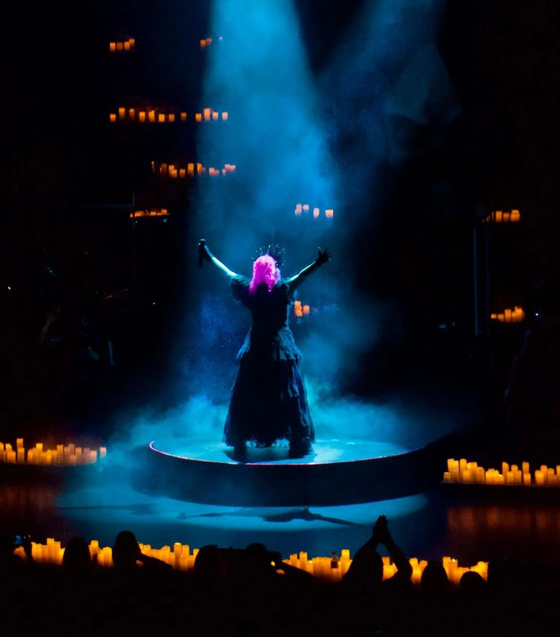 Performer with purple hair on a stage, arms raised, surrounded by blue lighting and fog, with candles surrounding the stage.