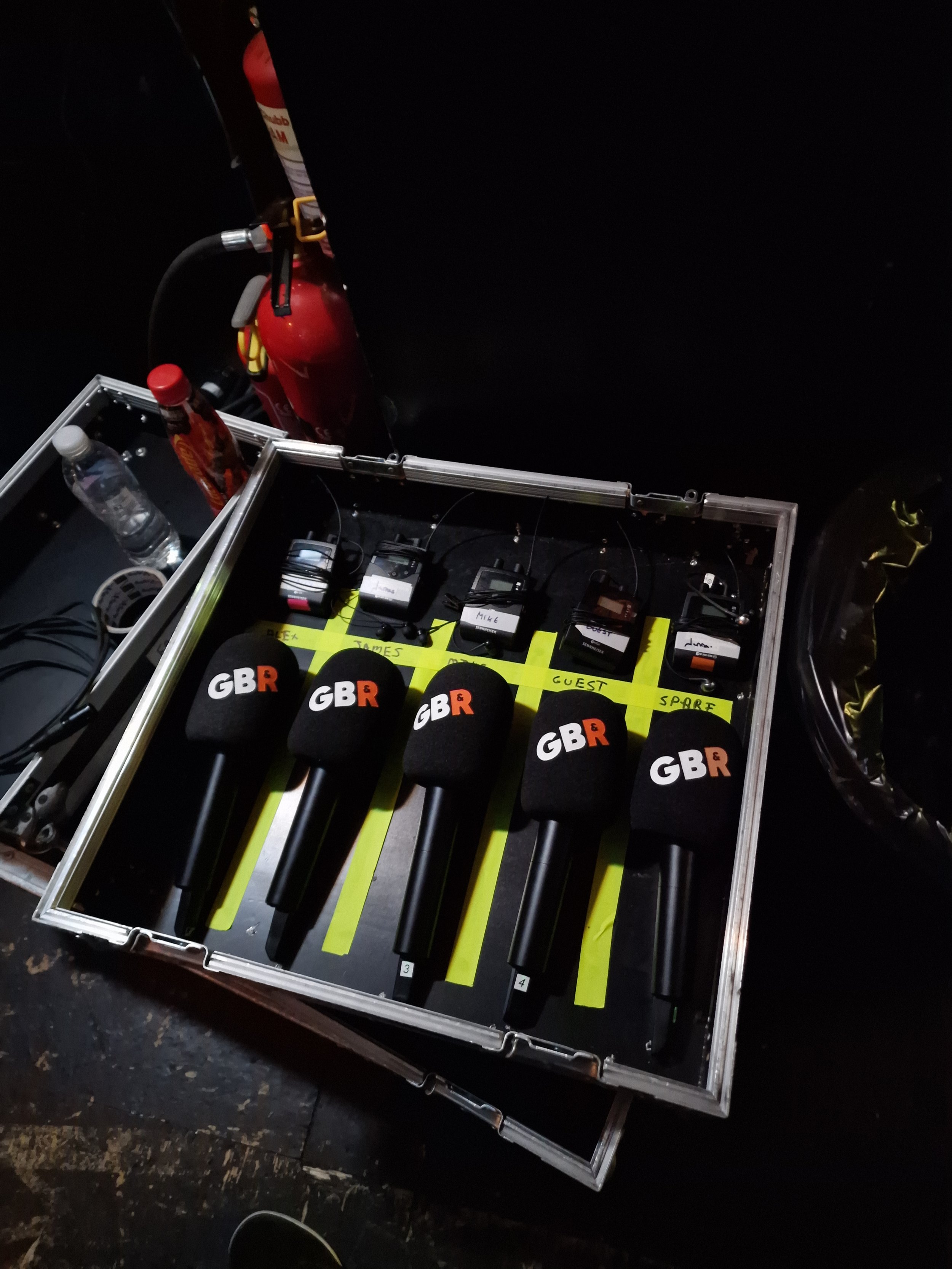 A collection of five microphones labeled GBR with foam covers, placed on a table. Behind them are five wireless receivers, each with a label for different speakers: Alex, James, Mike, Guest, and Spare. To the left, there are water bottles, a red fire