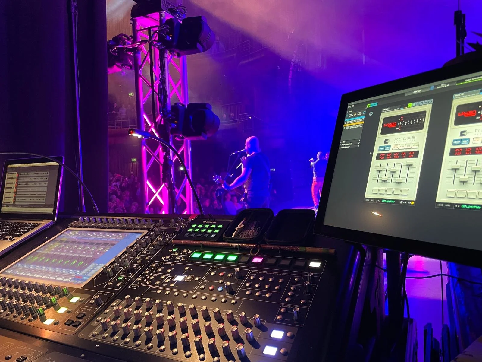 View from a sound mixing console overlooking a live music concert with purple and blue stage lighting and a performer singing with a guitarist.