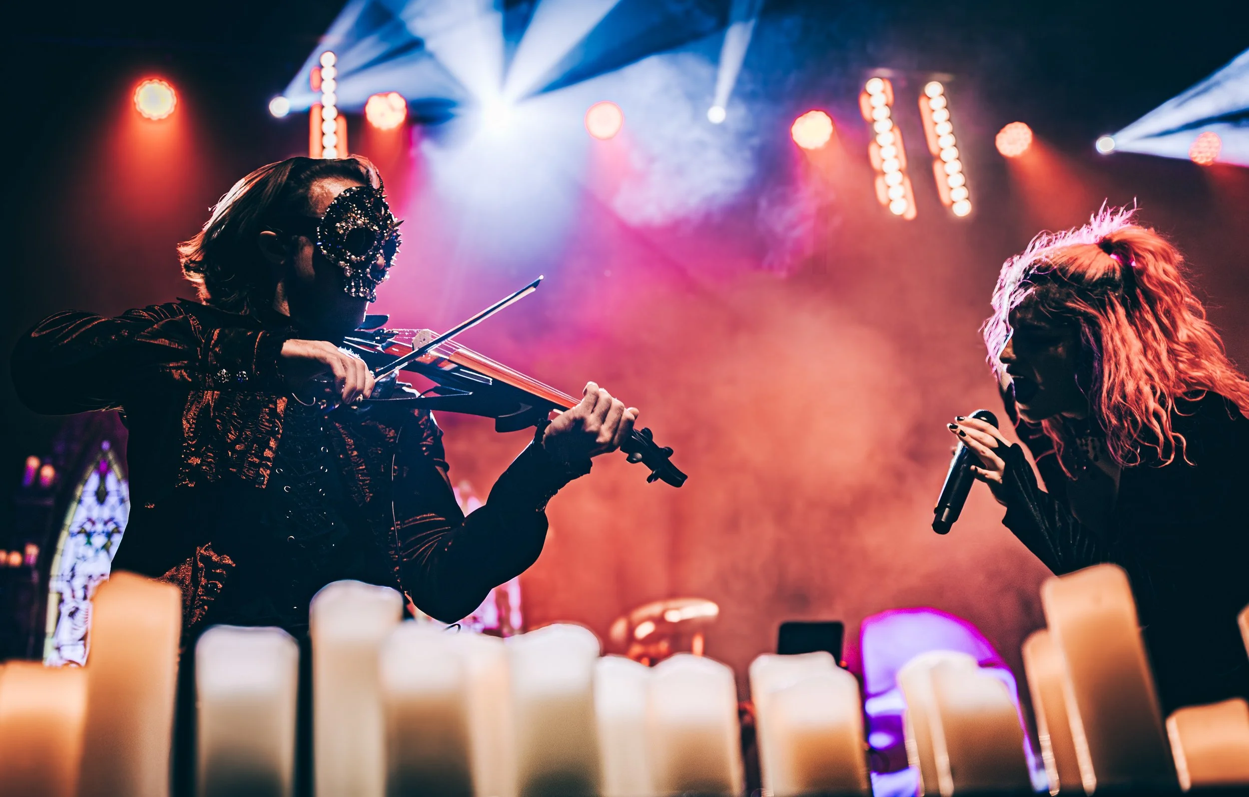 A musical performance featuring a violinist and a singer on stage with colorful, atmospheric lighting and candles in the foreground.