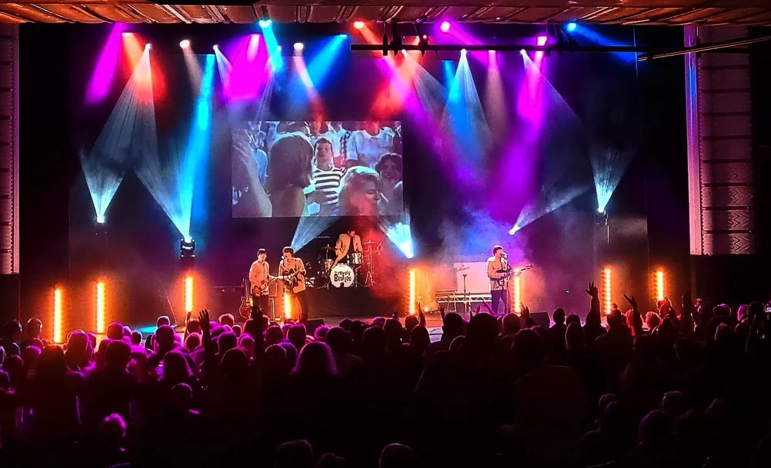 A lively concert stage with colorful lights and a band performing. Audience members are visible in the foreground, some raising their hands.