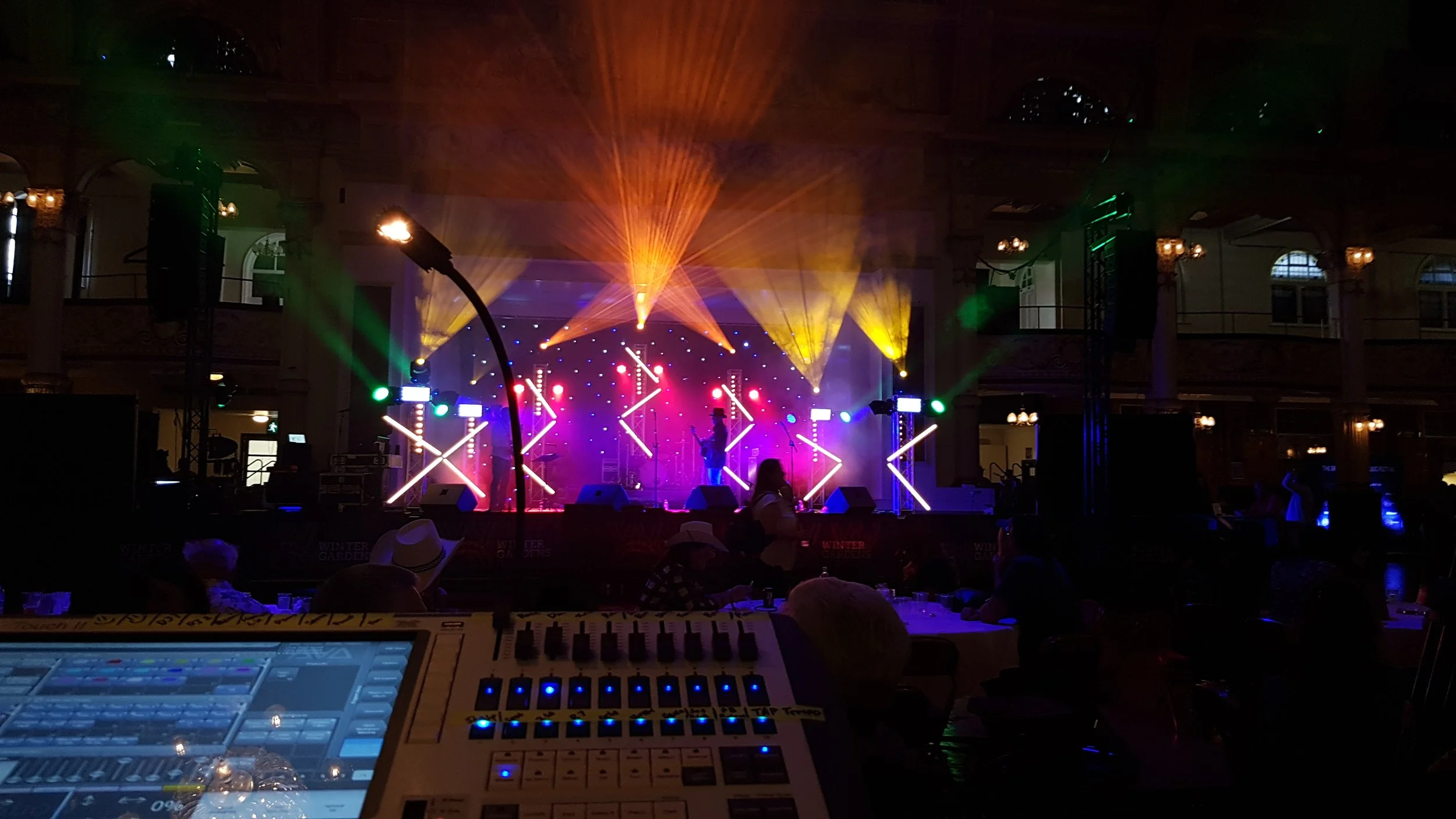 Stage with colorful lights and performer, audience seated in front, sound mixer in foreground.