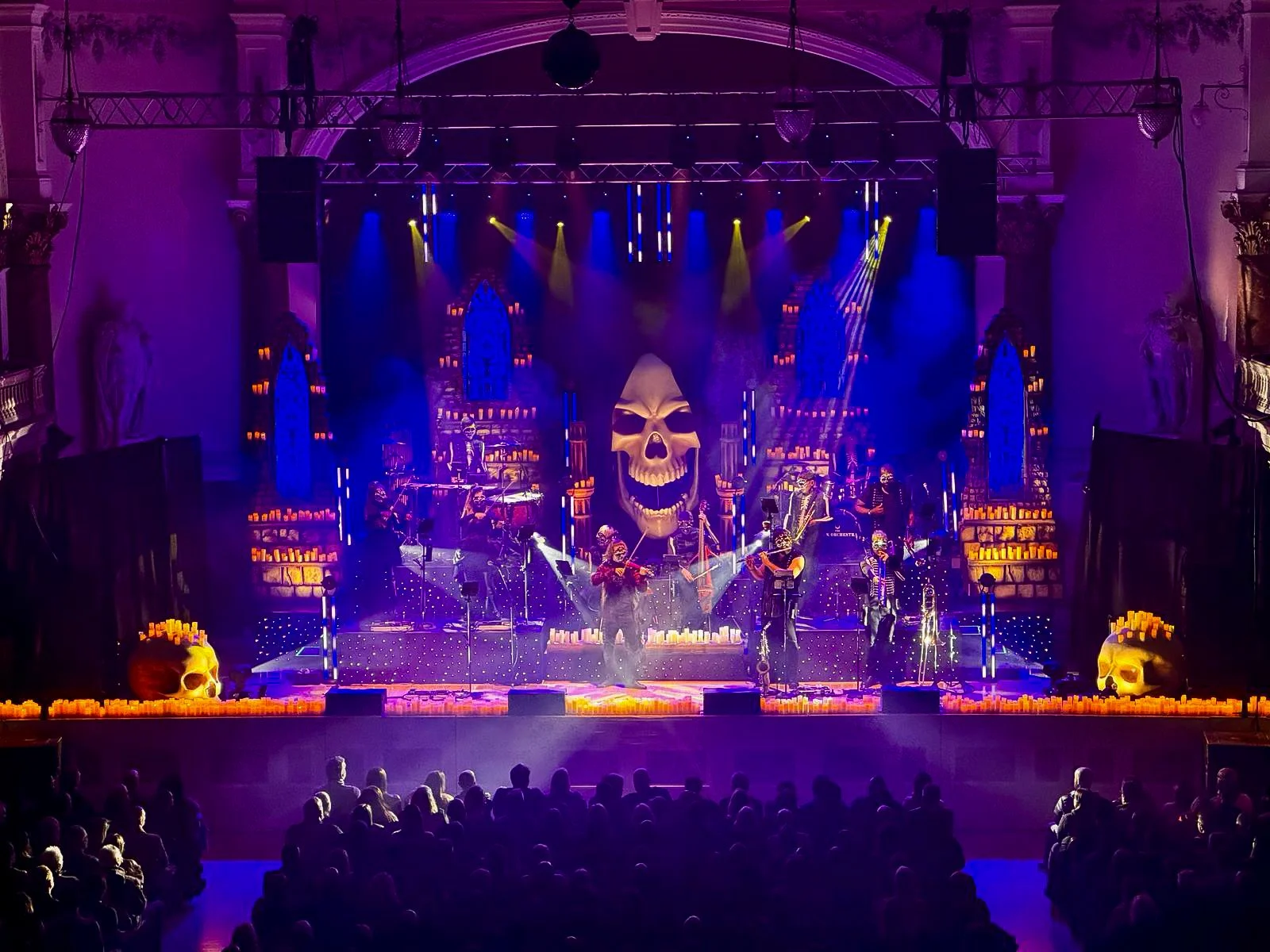 Concert stage with a large skull backdrop, candles, and Gothic architecture. Musicians are performing under purple and yellow lighting with candles on the edge of the stage.
