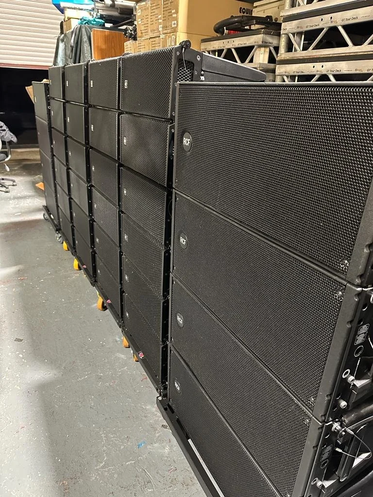A large array of black loudspeakers stacked in a storage room or warehouse.