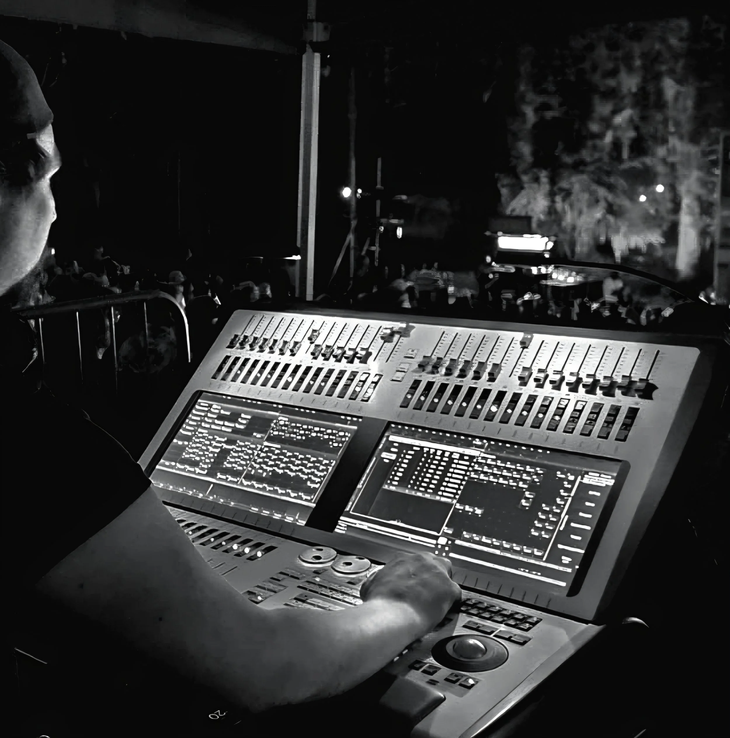 A person working at a DJ mixing console with multiple sliders and two digital screens, in a dark outdoor setting with a crowd and trees in the background.