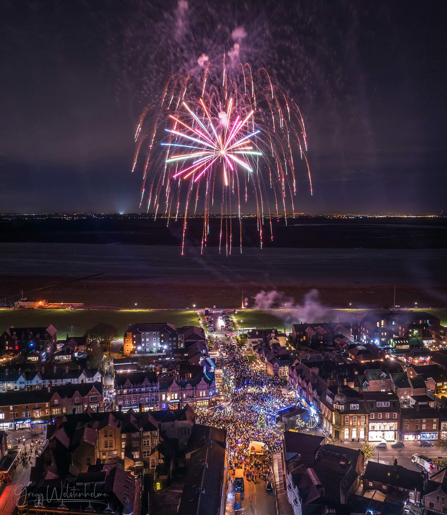 Nighttime scene of a fireworks display above a crowded street in a town or city, with houses, shops, and buildings illuminated below.