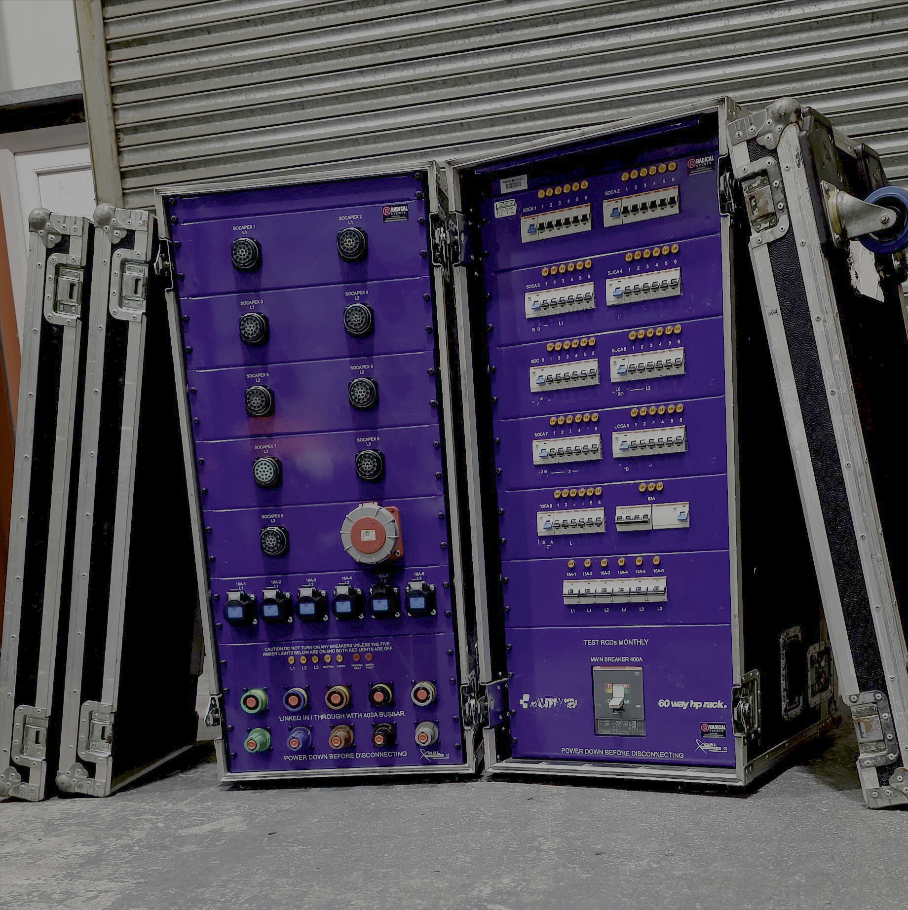 Two large purple electrical panels with various circuit breakers, switches, and connection ports, enclosed in metal frames, leaning against a metal shutter door.