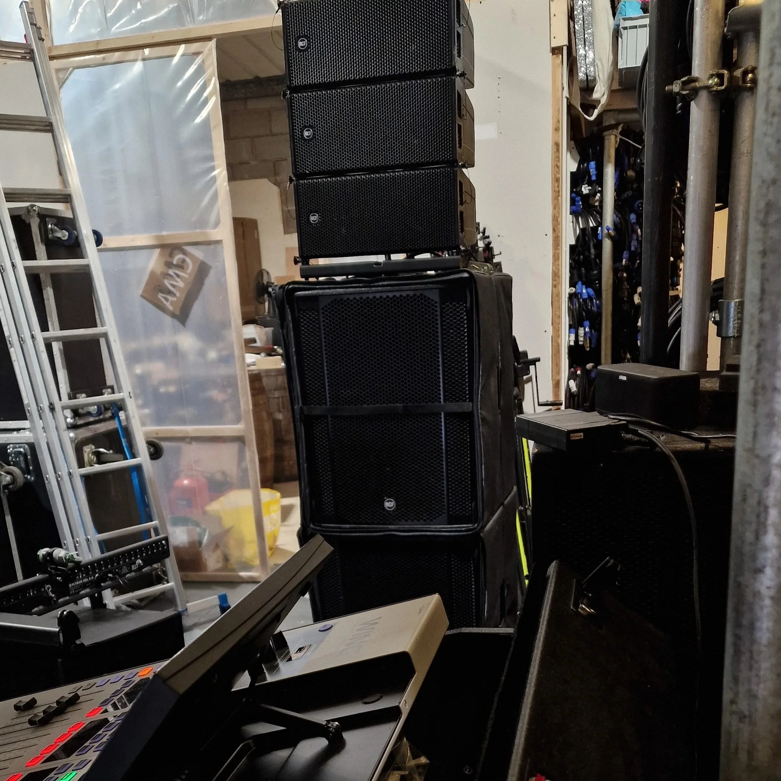 Stack of six black loudspeakers on a stage or workspace, with equipment and tools visible around.