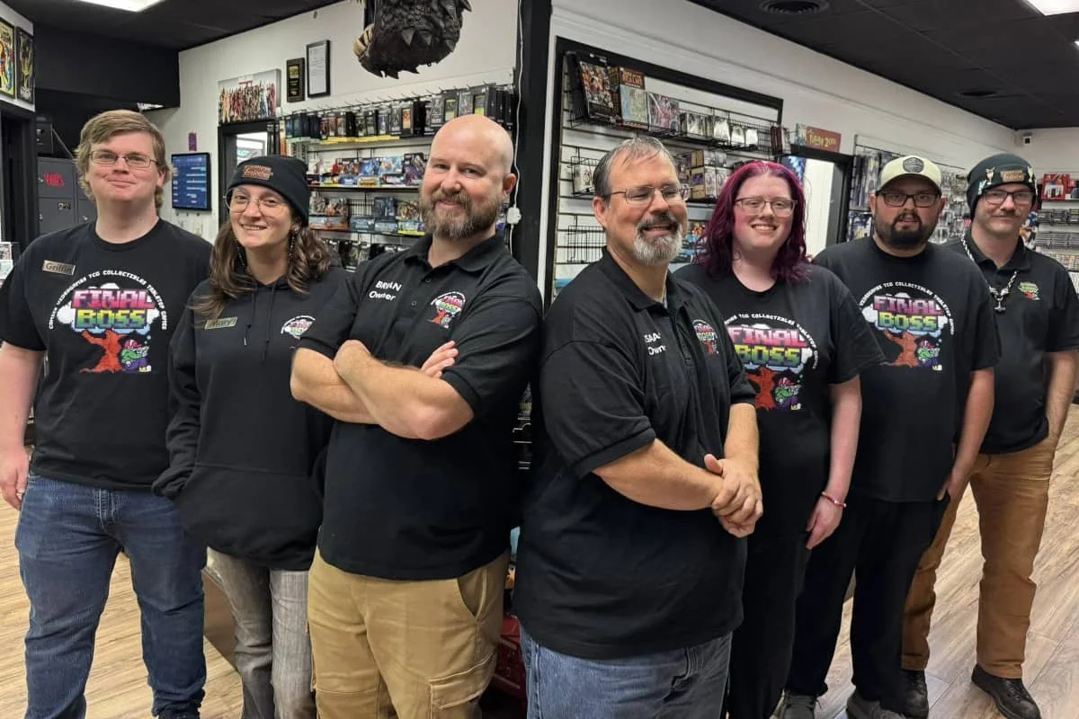 Group of seven people standing in a store with shelves of collectibles and merchandise behind them. They are smiling and wearing black shirts, some with the 'Final Boss' logo.