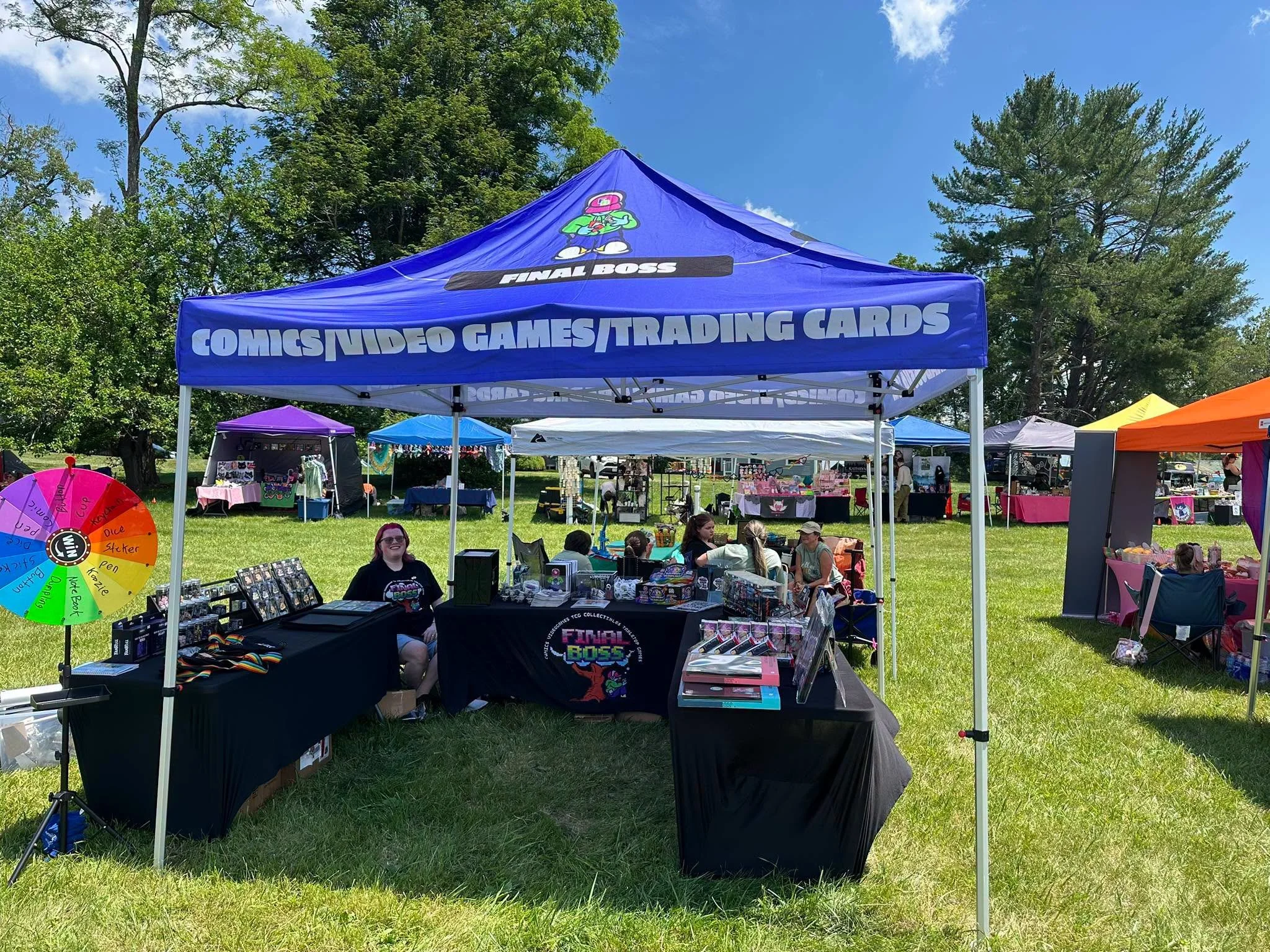 Outdoor vendor booth with a blue canopy tent labeled 'Comics Video Games Trading Cards' and a smaller purple tent behind it, at a sunny park fair with trees and people.