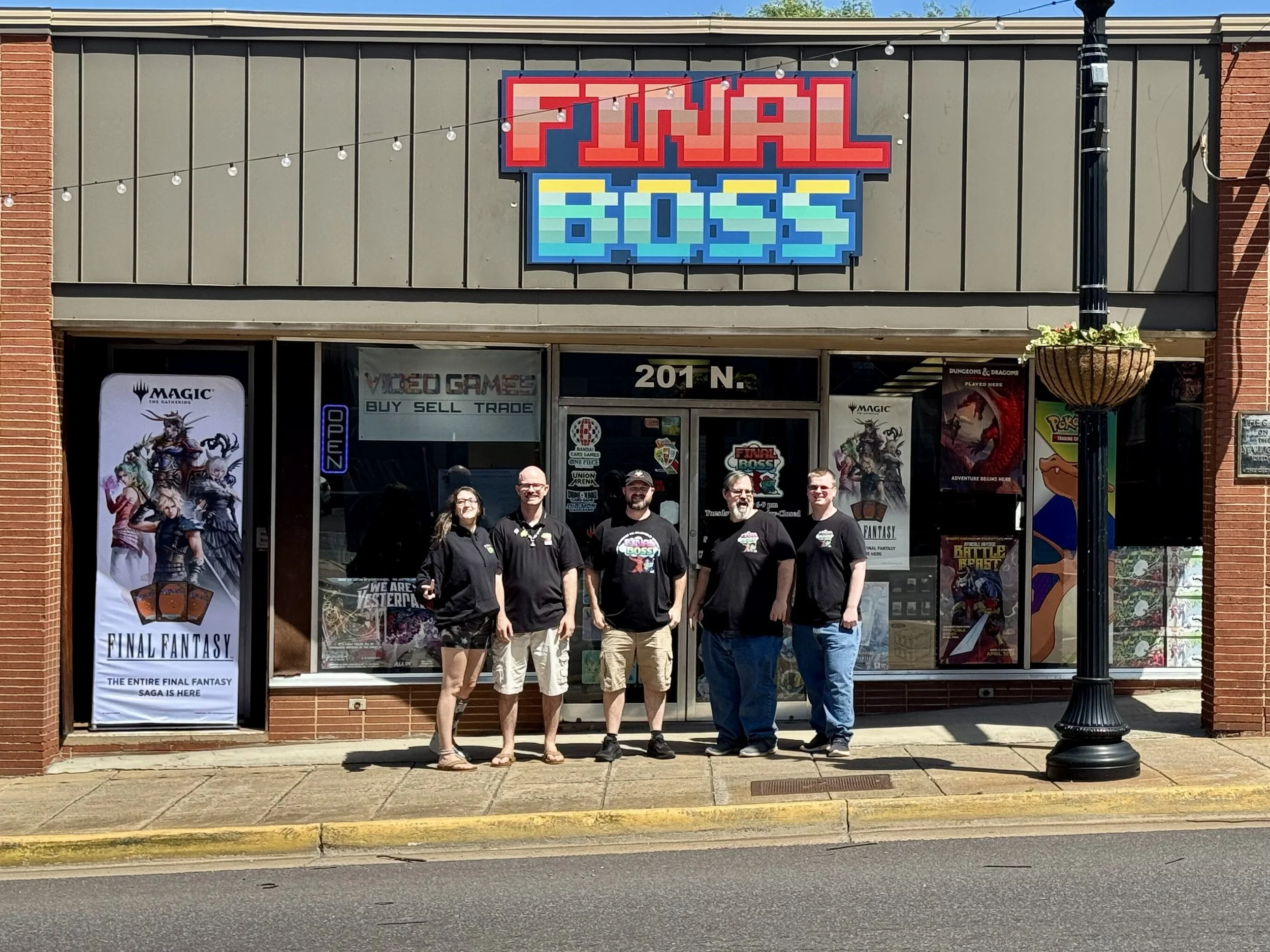 Group of five people standing outside gaming store with sign for 'Final Fantasy' and promotional posters in window.