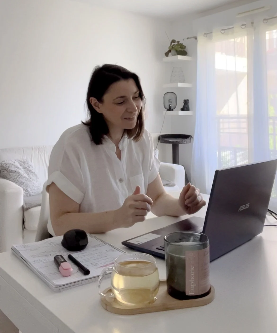 Une femme assise à une table de cuisine, utilisant un ordinateur portable, avec un carnet, un stylo, une souris, un thermos, une tasse de thé, et une bougie allumée sur la table.