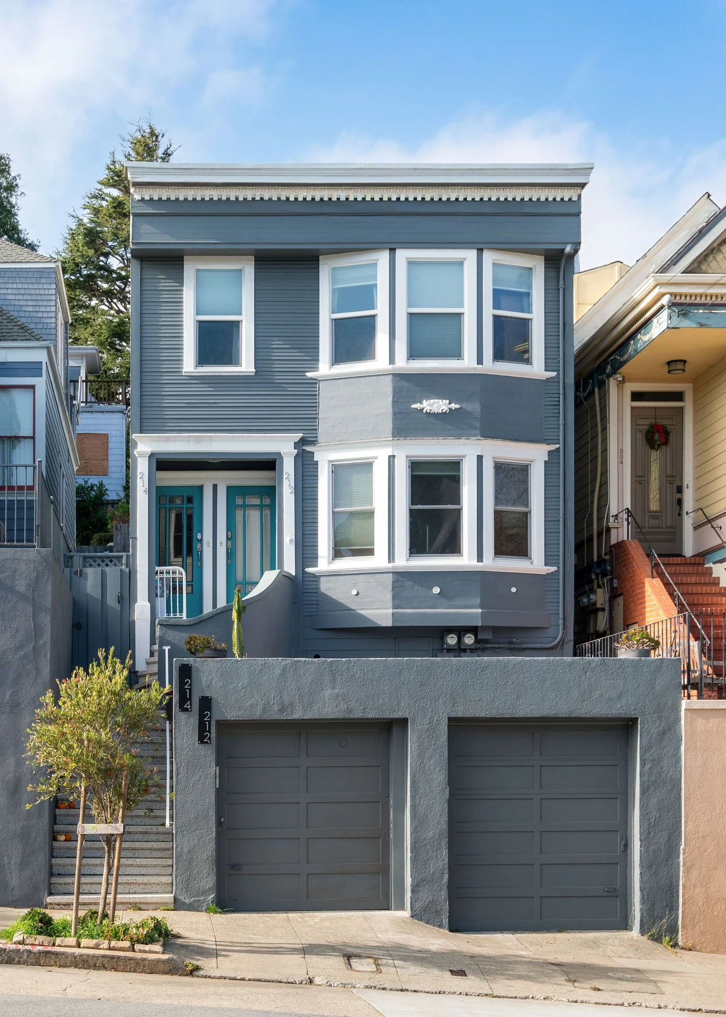A multi-story residential house painted in blue with white trim, featuring two garages and multiple large windows, with neighboring homes visible on either side and a clear sky above.