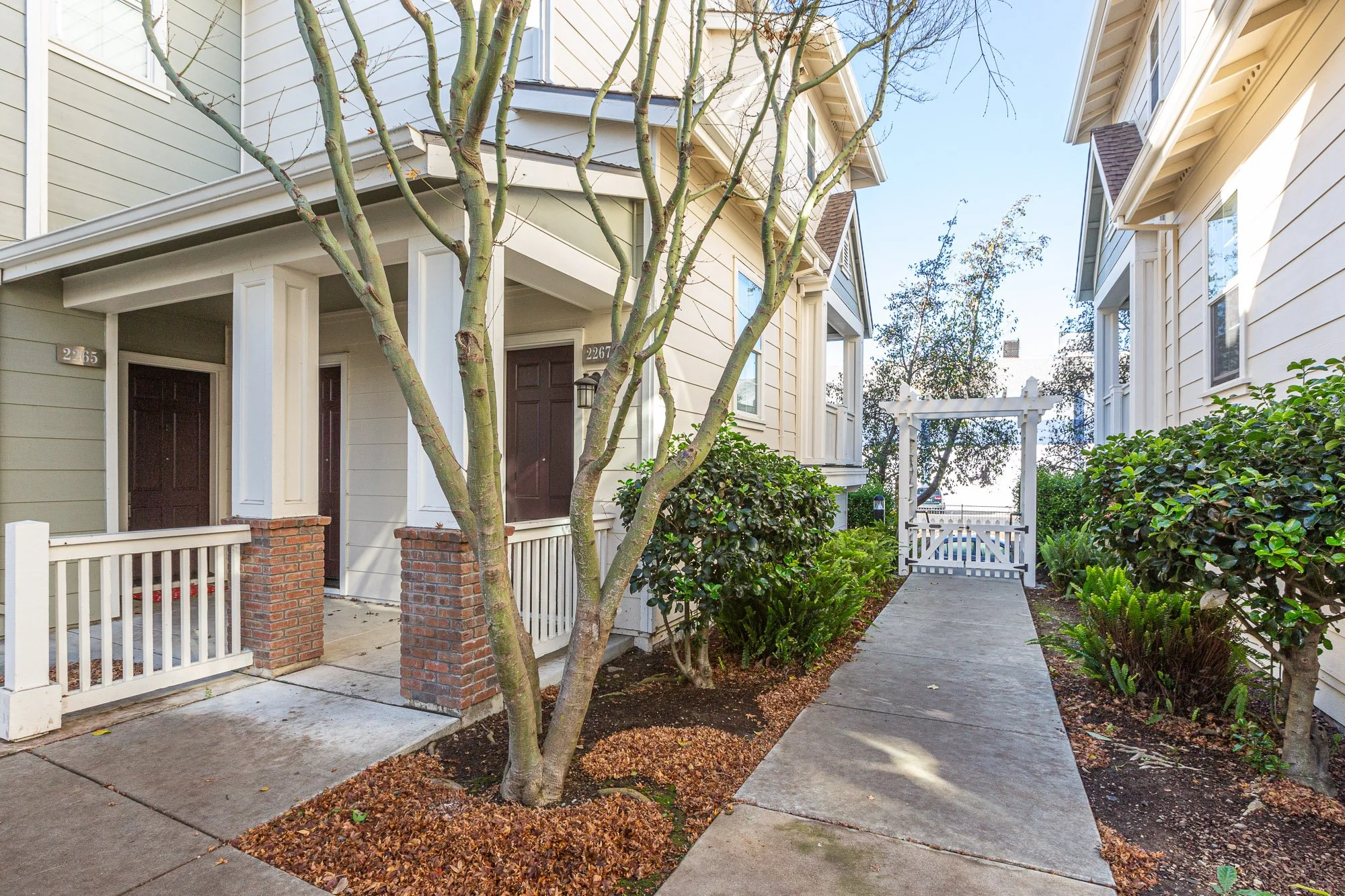 A residential apartment complex with beige siding, black front doors, a sidewalk, a white gate, trees, and shrubs in the front yard.