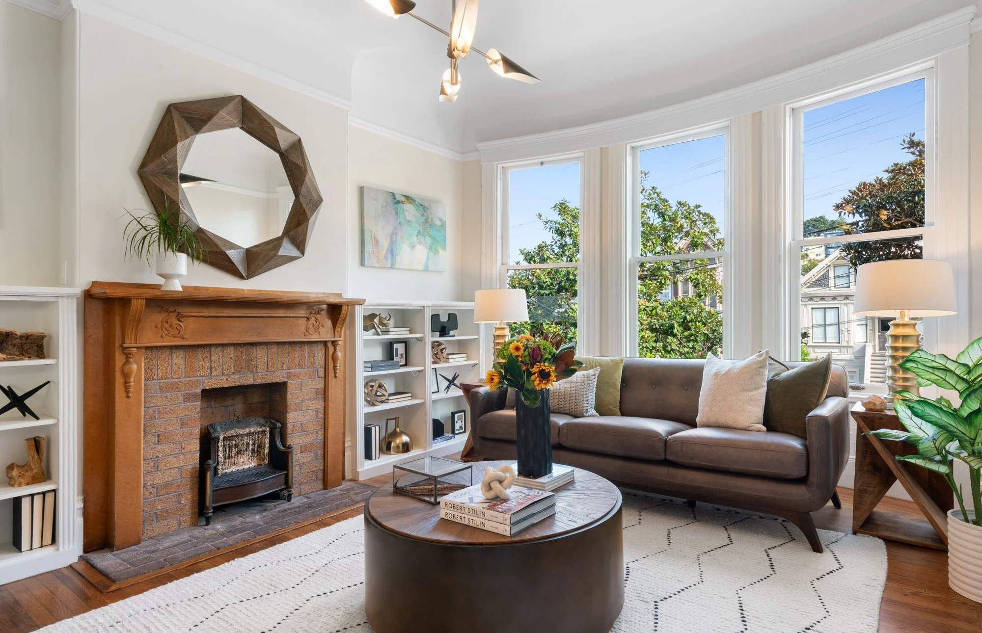 Living room with a brick fireplace, wooden mantel, large windows, sofa with cushions, side table, lamp, bookshelf, and colorful flower vase.