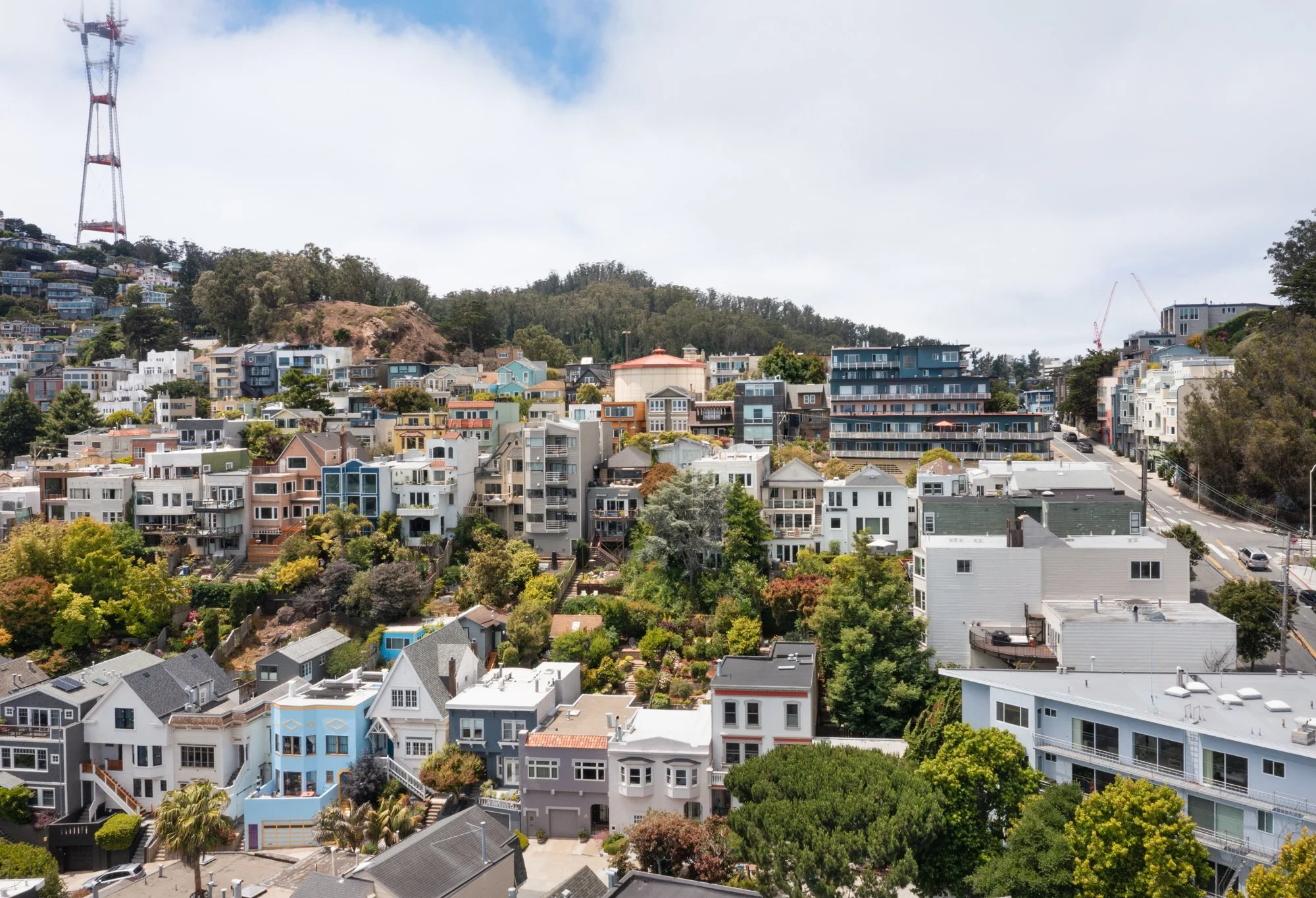 Hilly neighborhood with a variety of colorful modern houses, trees, and a communication tower on a partly cloudy day.