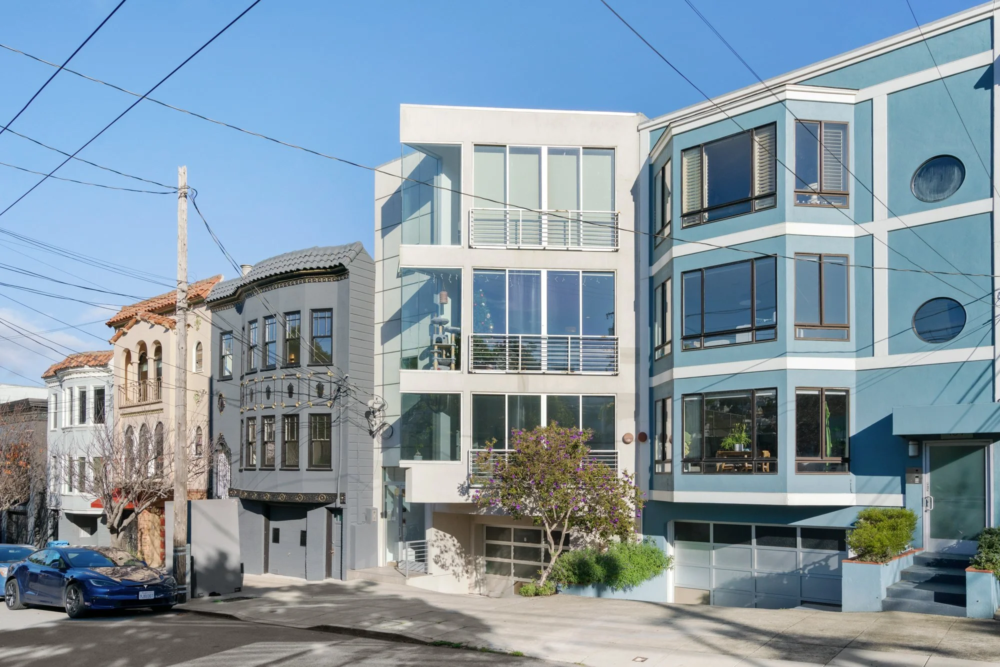 Multiple modern multi-story residential buildings with large windows along a street under a clear blue sky, with power lines overhead and parked cars.