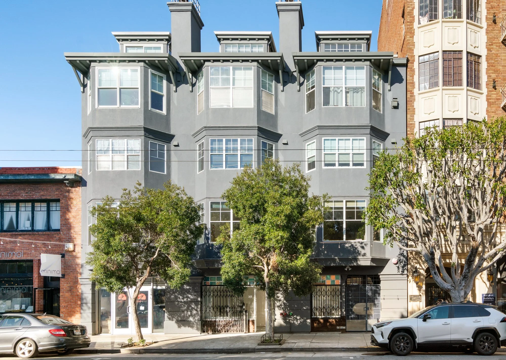 A modern, gray multi-story building with large windows and a gated entrance, flanked by two trees on the sidewalk, with parked cars in front.