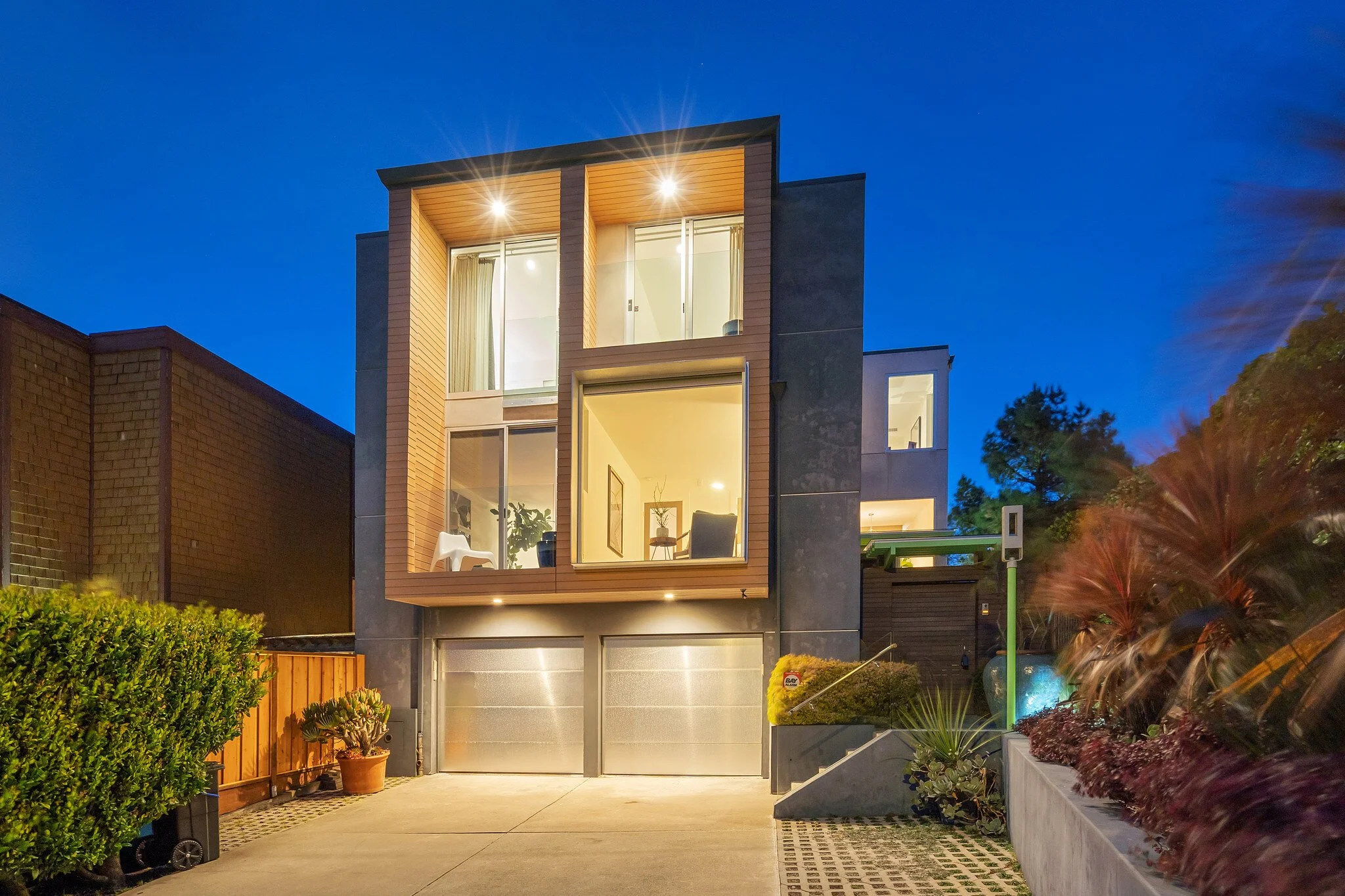 Modern multi-story house with illuminated interior, large glass windows, and a driveway at dusk.
