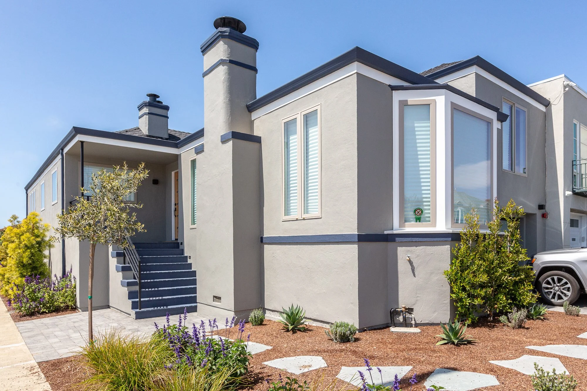 Modern two-story house with a light gray exterior, multiple large windows, and a front door accessible via gray painted stairs. The landscaped front yard features small trees, shrubs, and purple flowers, with a concrete pathway. There is a parked SUV on the right side.