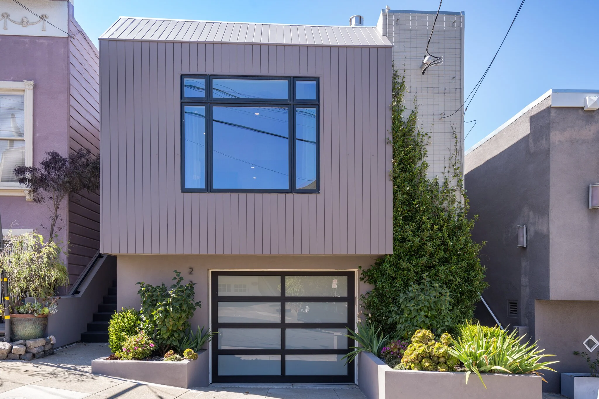 Modern two-story house with gray exterior, large front-facing window, black garage door, staircase on the left, and landscaped front yard with various plants and flowers.