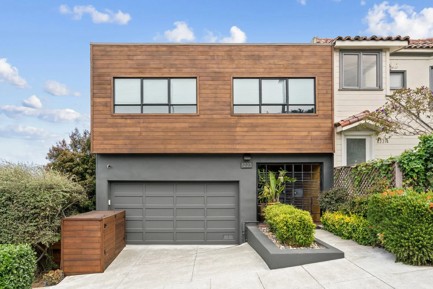 Modern two-story house with a dark gray garage door, a wooden upper facade, and a small front garden with green bushes and a path leading to the entrance.
