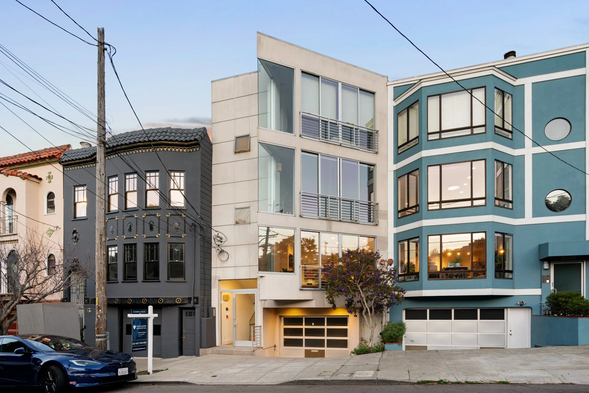 A row of modern and traditional style residential buildings on a sunny street, with a black car parked in front and power lines overhead.