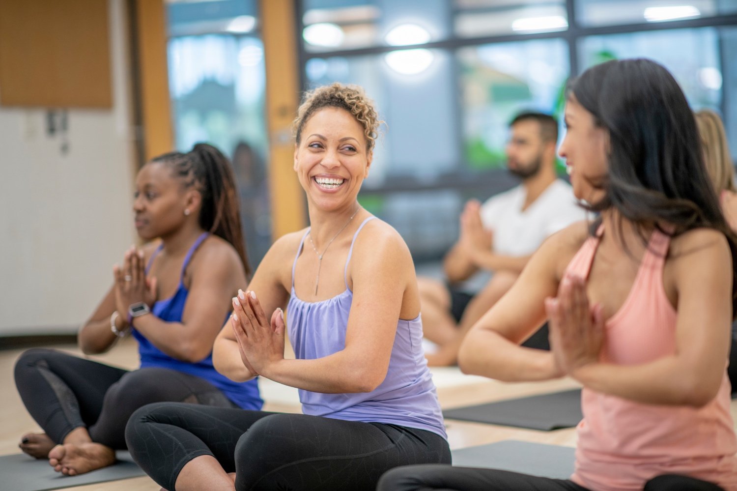Group of people practicing yoga or meditation in a studio, sitting cross-legged with hands in prayer position, some smiling.