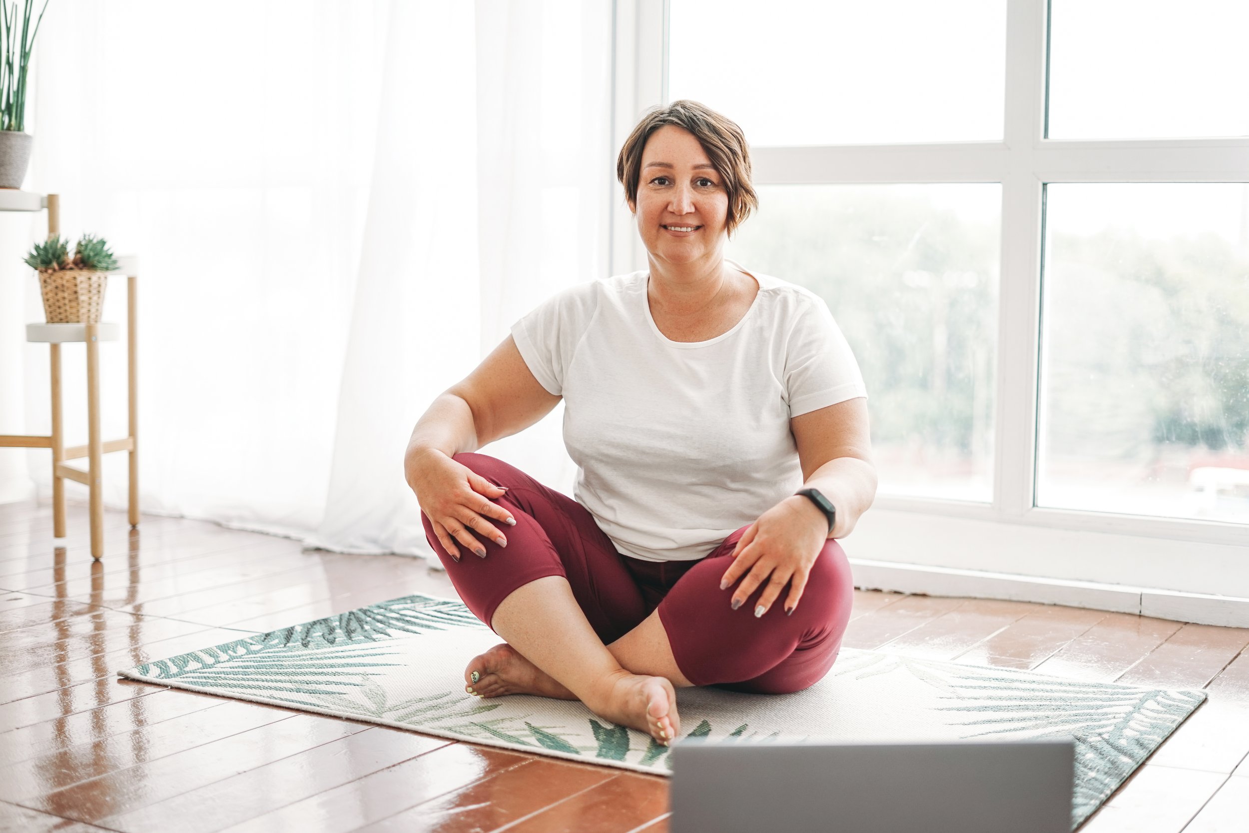 Woman sitting cross-legged on a yoga mat in a bright room with large windows, smiling at the camera